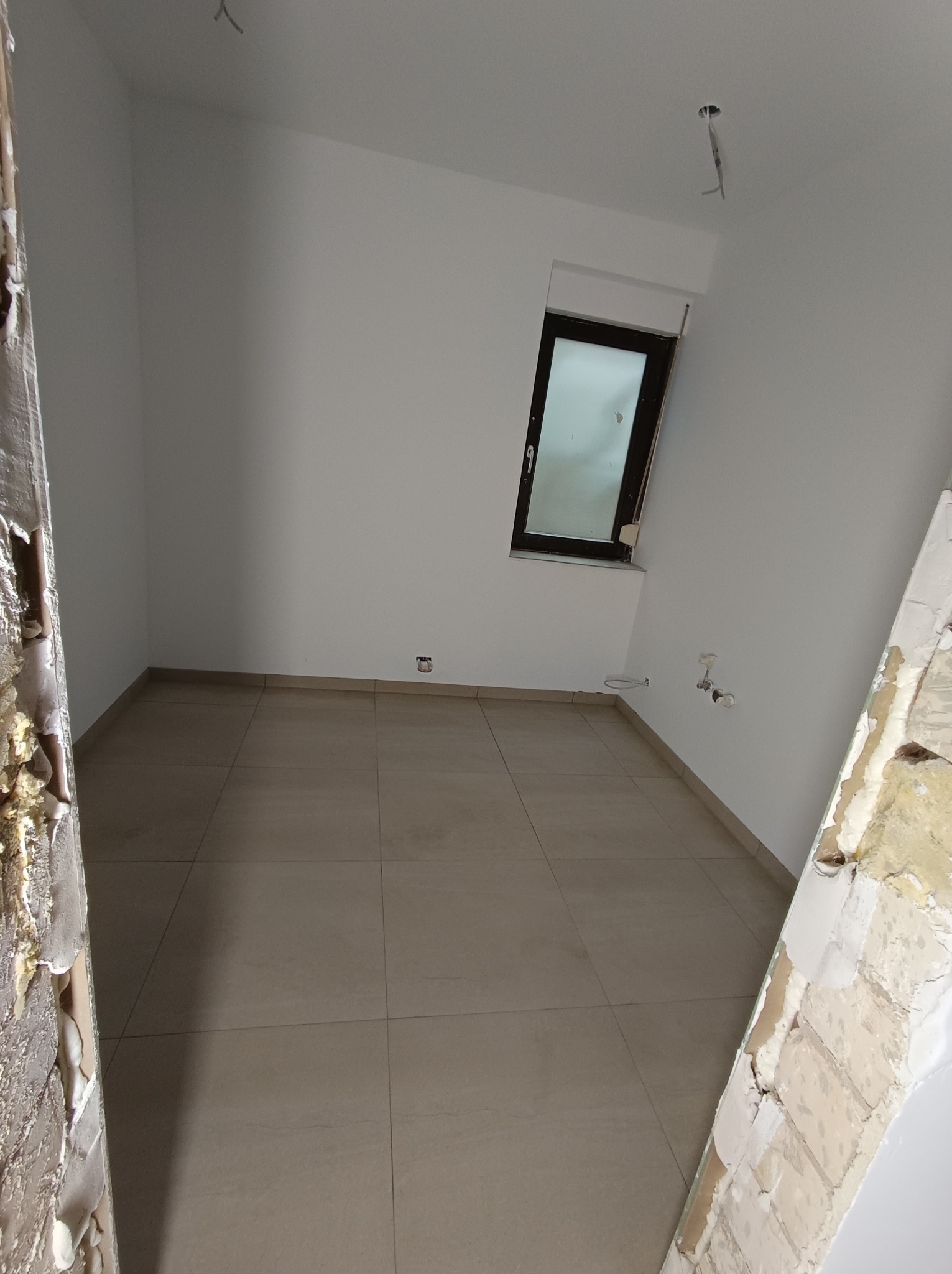 Pusty pokój w trakcie remontu, z nowymi płytkami na podłodze, świeżo pomalowanymi ścianami, odsłoniętymi przewodami elektrycznymi na suficie i ościeżnicami w stanie surowym.