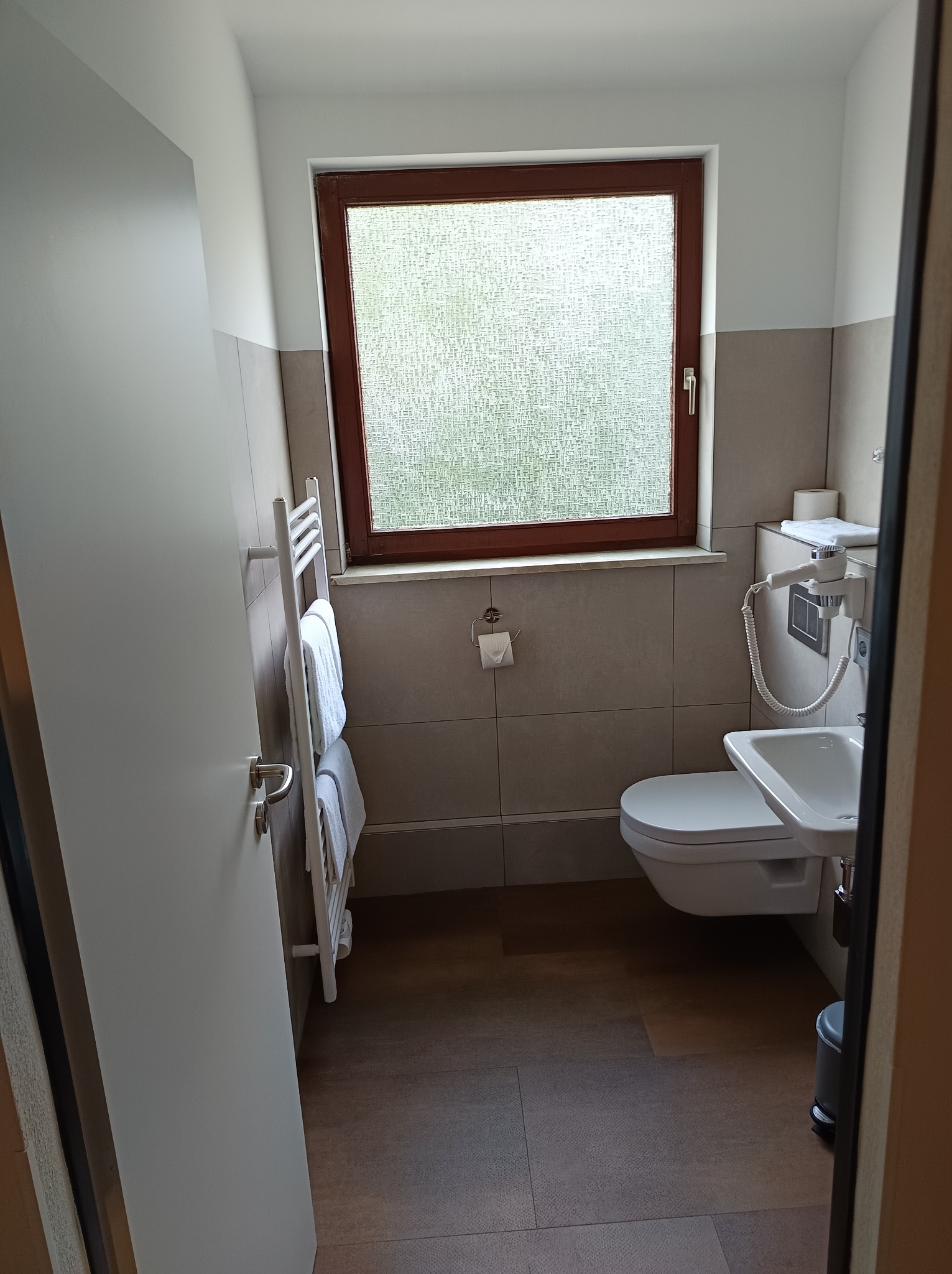 Nowoczesna łazienka po remoncie: biała ceramika, wisząca umywalka, grzejnik drabinkowy z ręcznikami, okno z matową szybą, brązowe płytki podłogowe.