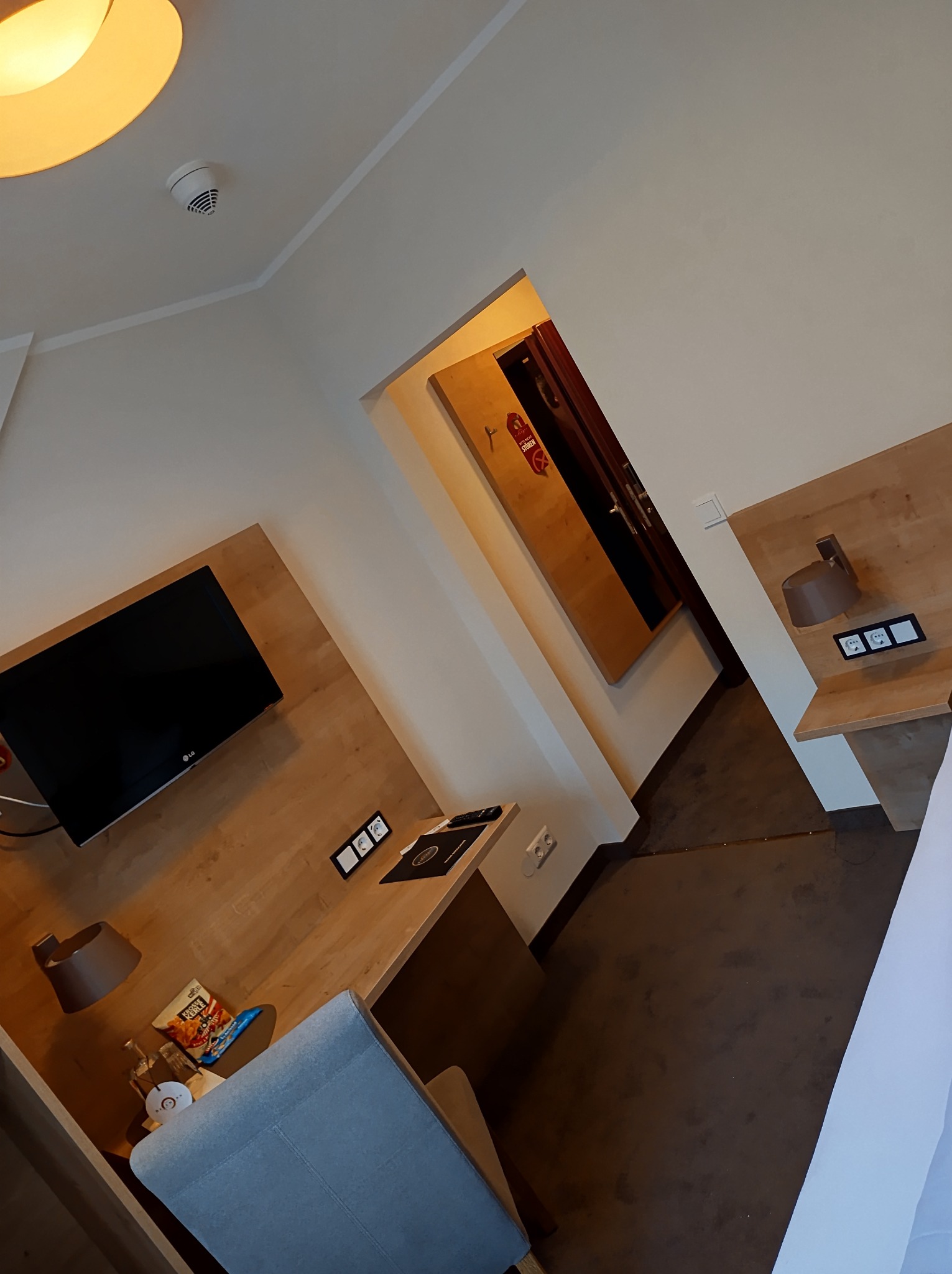 Wnętrze pokoju hotelowego z drewnianą zabudową, telewizorem, biurkiem z lampką, krzesłem i otwartymi drzwiami do garderoby.