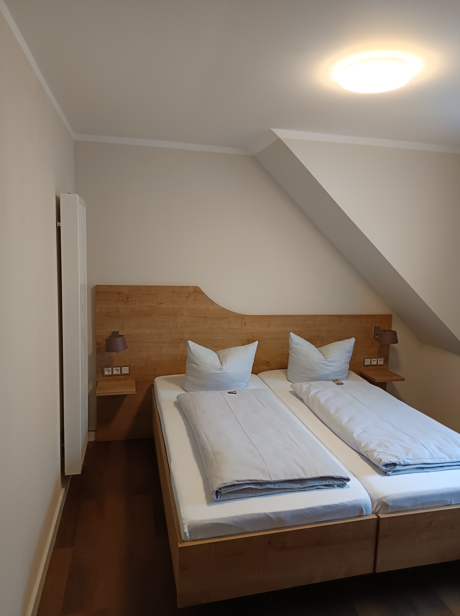 Pokój z dwoma pojedynczymi łóżkami z drewnianymi ramami i zagłówkiem, szafkami nocnymi z lampkami, poddasze z oknem dachowym, białe ściany i brązowa podłoga.