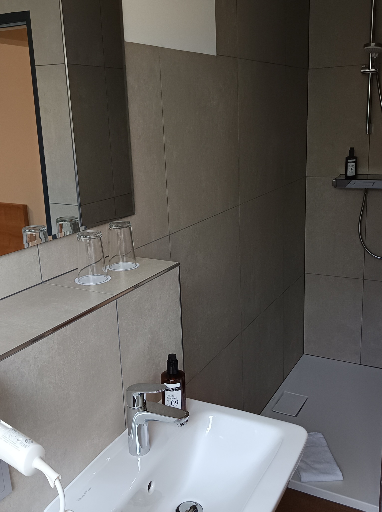 Nowoczesna łazienka z umywalką Villeroy & Boch, baterią chromowaną, szklankami na blacie i kabiną prysznicową z odpływem liniowym, wykończona płytkami w odcieniach szarości.