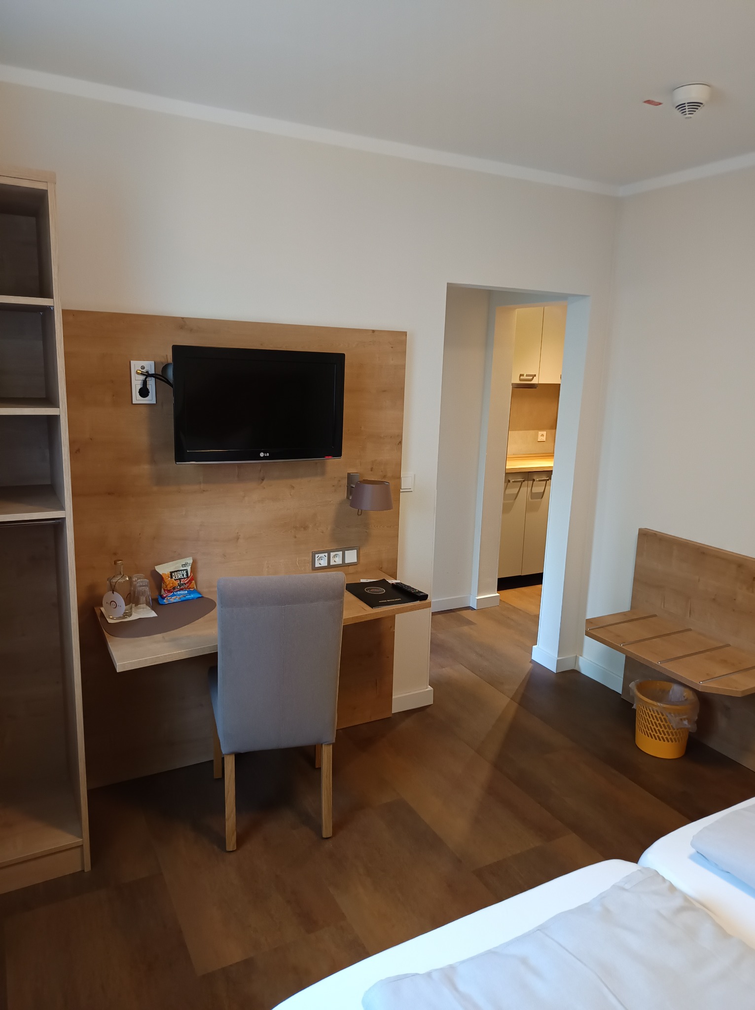 Wnętrze pokoju hotelowego z drewnianą podłogą, biurkiem z telewizorem LG, szarą krzesłem, szafą i widokiem na aneks kuchenny.