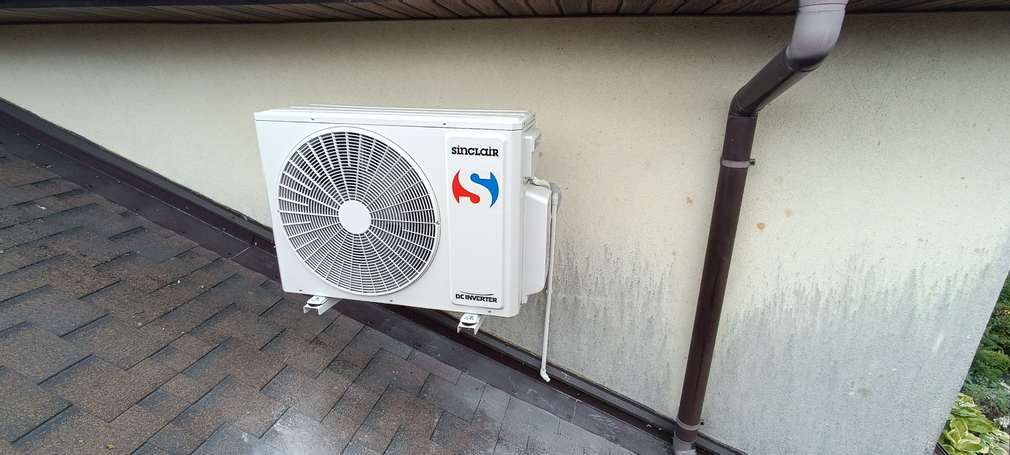 Zewnętrzna jednostka klimatyzacji Sinclair DC Inverter zamontowana na ścianie domu, widoczny brązowy system rynnowy i dach z brązowej dachówki bitumicznej.