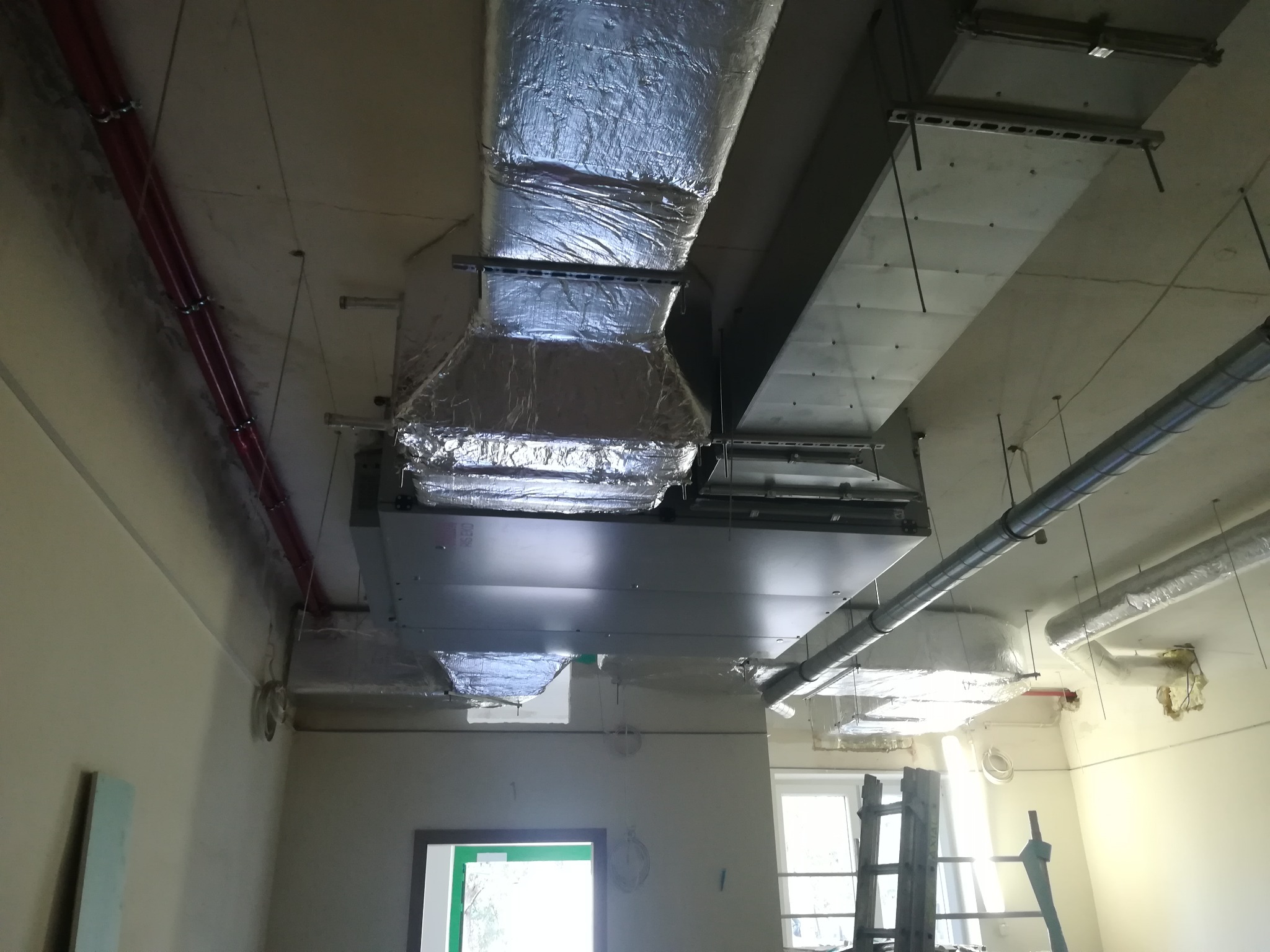 Widok na sufit w trakcie montażu instalacji wentylacyjnej z widocznymi kanałami izolowanymi folią aluminiową, elementami mocującymi oraz fragmentem drabiny opartą o ścianę z oknem.