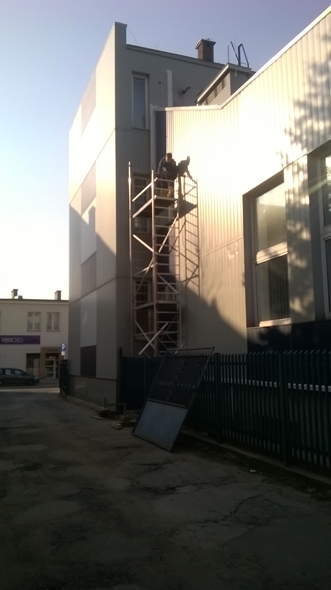 Dwóch pracowników na rusztowaniu przy ścianie budynku komercyjnego, prawdopodobnie montaż lub serwis instalacji. Widoczny fragment elewacji, okna i błękitne niebo w tle.