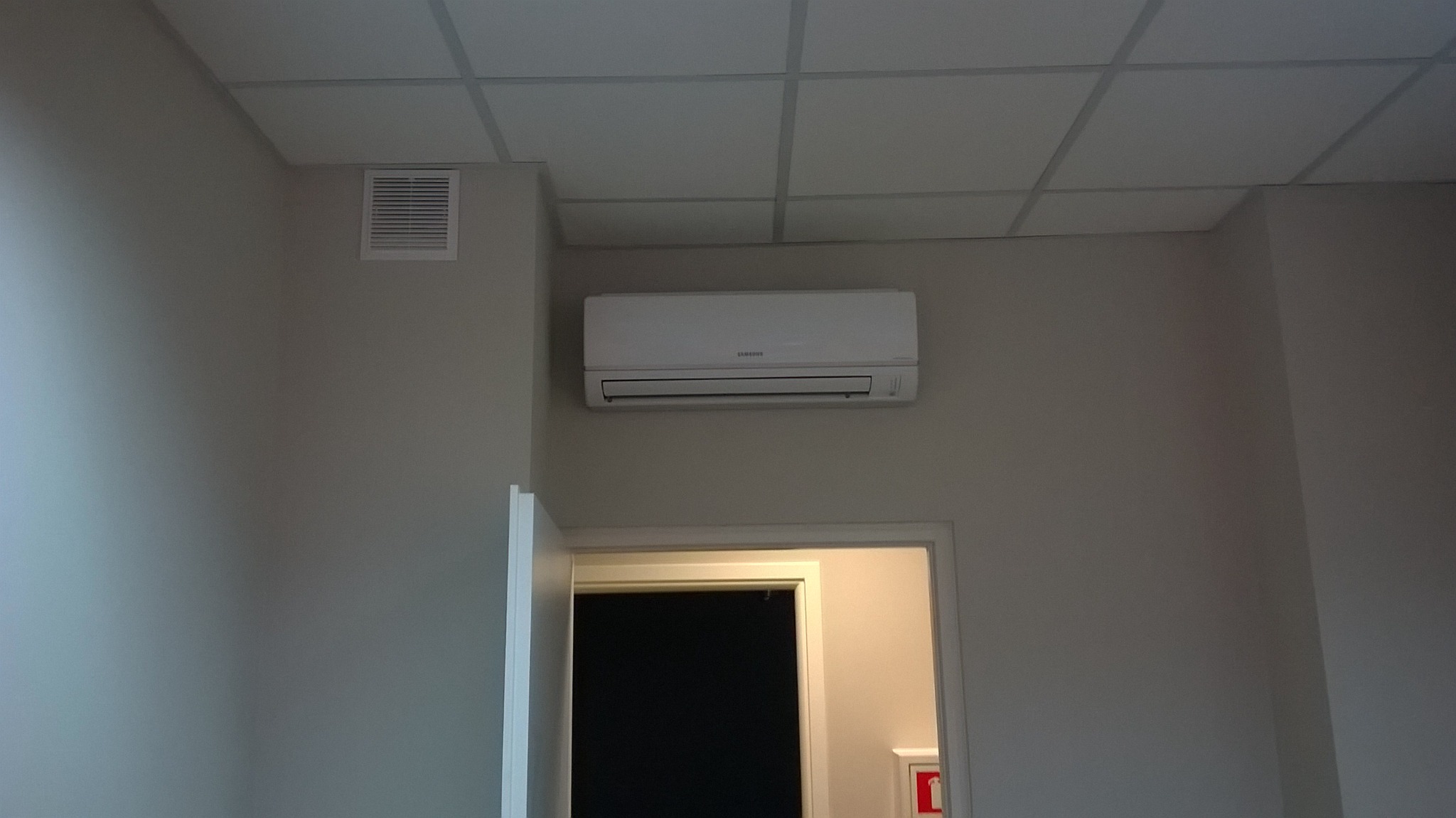 Biały klimatyzator Samsung zamontowany na ścianie nad otwartymi drzwiami w pomieszczeniu z podwieszanym sufitem kasetonowym i kratką wentylacyjną.