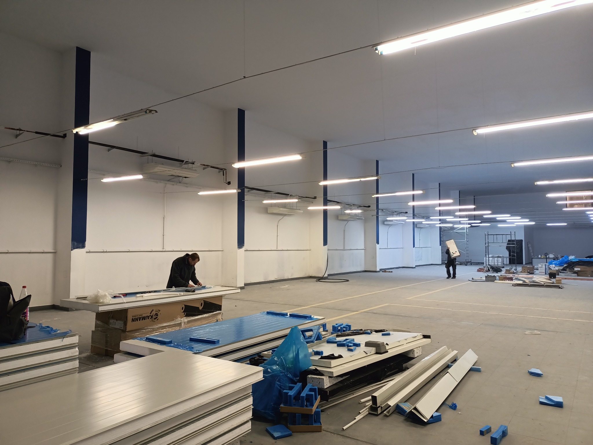 Duża hala w trakcie montażu paneli izolacyjnych, widoczne elementy instalacji wentylacyjnej pod sufitem, pracownik przycina panel na stole roboczym, w tle osoba przenosi karton.