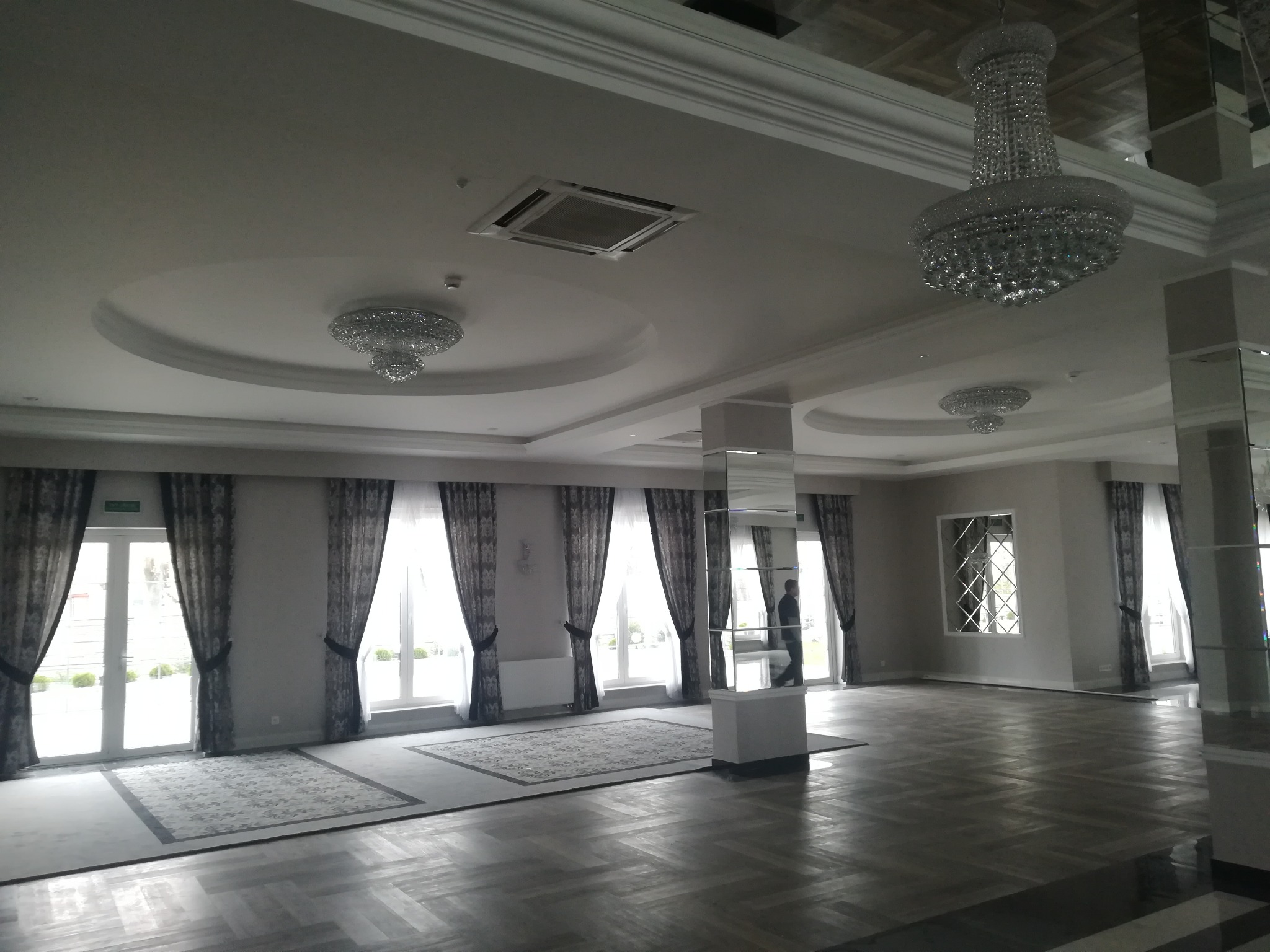 Wnętrze sali balowej z parkietem w jodełkę, żyrandolami kryształowymi, dużymi oknami z zasłonami i kratką wentylacyjną w suficie podwieszanym.