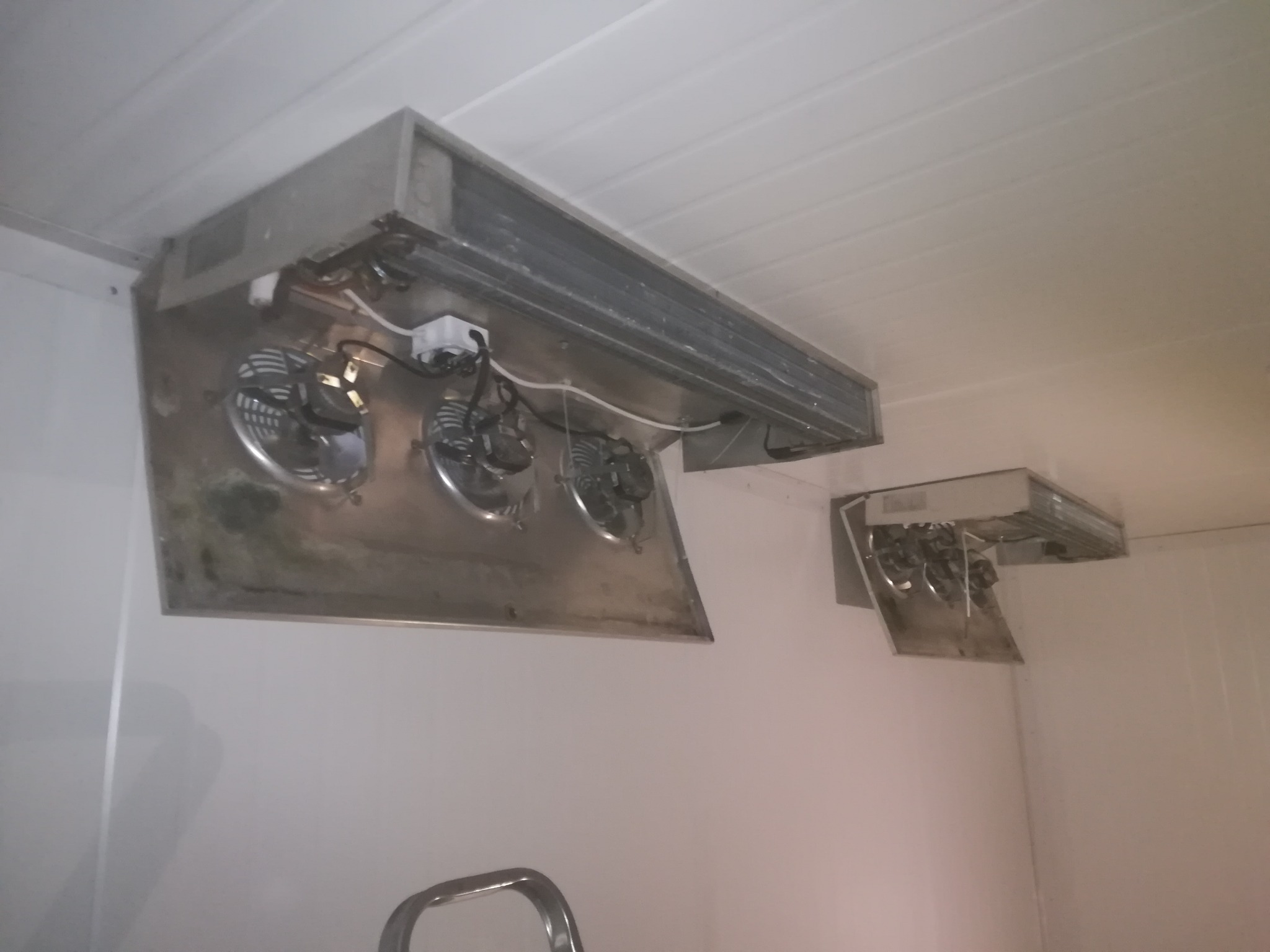 Dwa przemysłowe klimatyzatory sufitowe z trzema wentylatorami każdy, zamontowane w chłodni, widoczne przewody elektryczne i ślady użytkowania na obudowach.