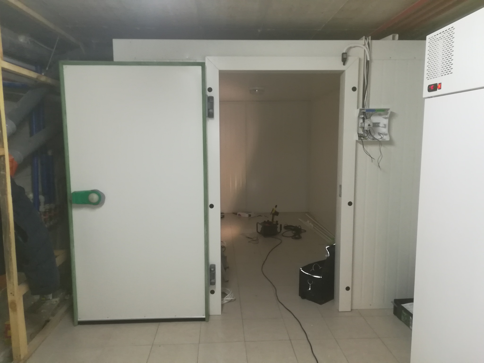 Wnętrze chłodni z otwartymi drzwiami, widoczne narzędzia i materiały budowlane wewnątrz, boczna ściana z widoczną instalacją elektryczną i agregatem chłodniczym.