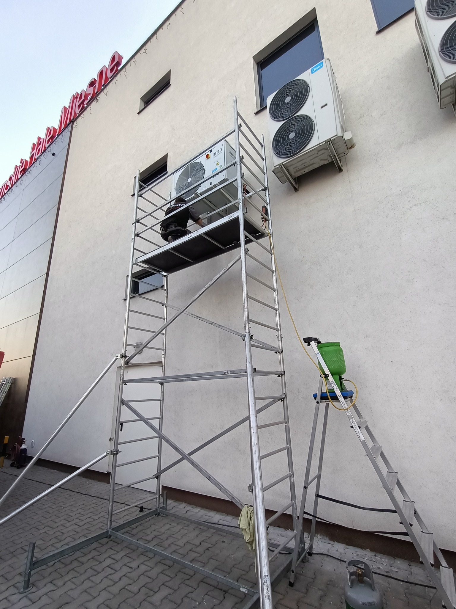 Pracownik serwisu na rusztowaniu przy zewnętrznej jednostce klimatyzacji budynku komercyjnego, obok drabina z zielonym pojemnikiem i butla z gazem.