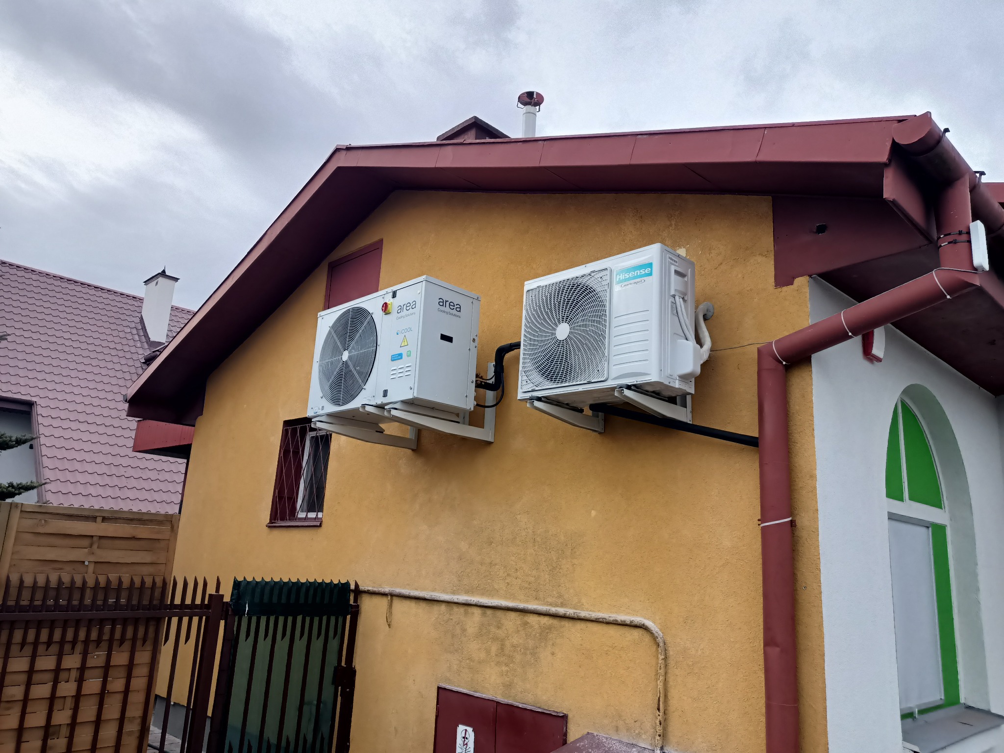 Dwa klimatyzatory różnych marek (Area i Hisense) zamontowane na elewacji domu z żółtym tynkiem, widoczne okno z kratą i fragment ogrodzenia.