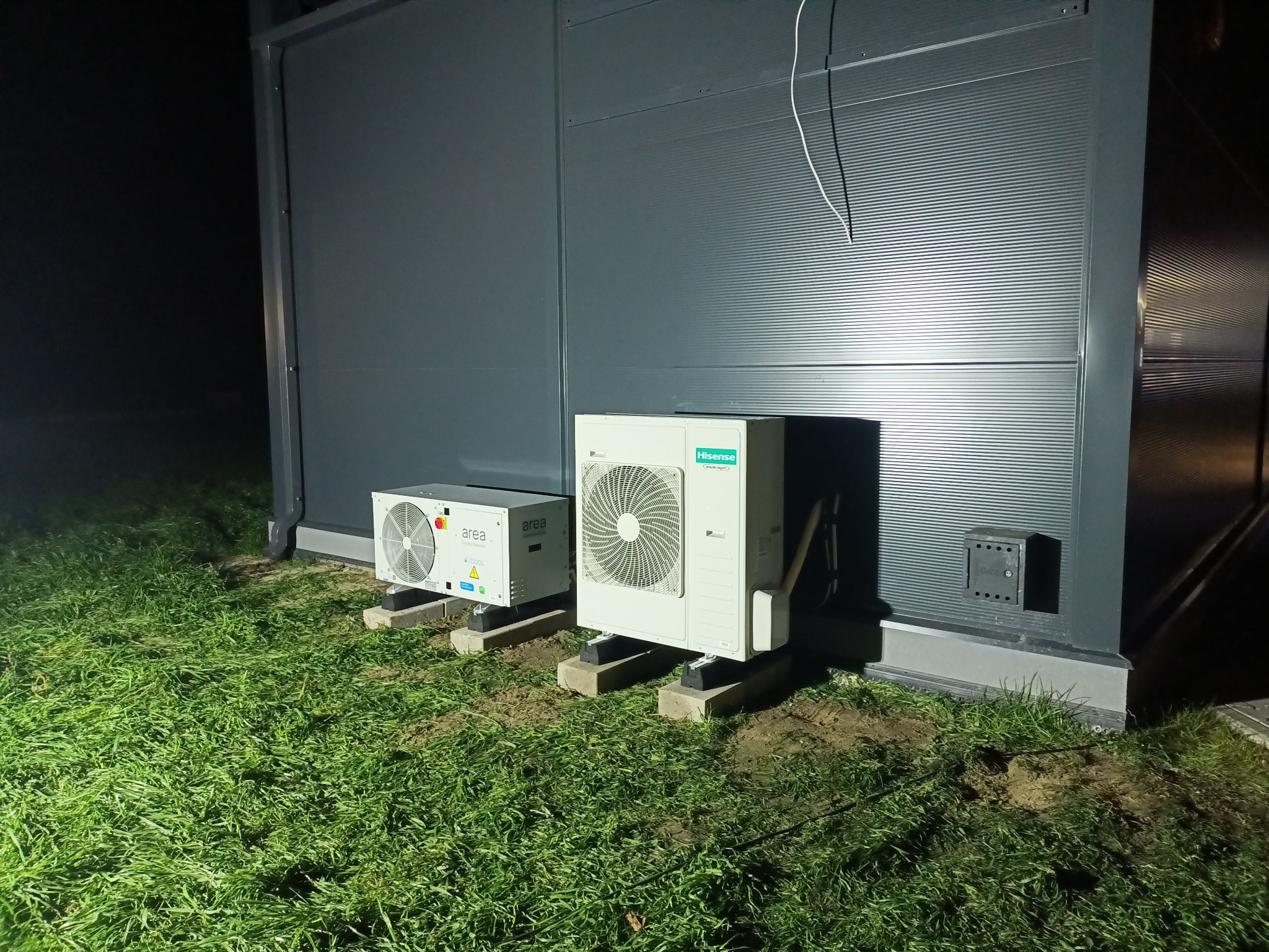 Dwie jednostki zewnętrzne klimatyzacji (Hisense i Area) zamontowane na betonowych podstawach przy szarej ścianie budynku w trawie, widoczne przewody.