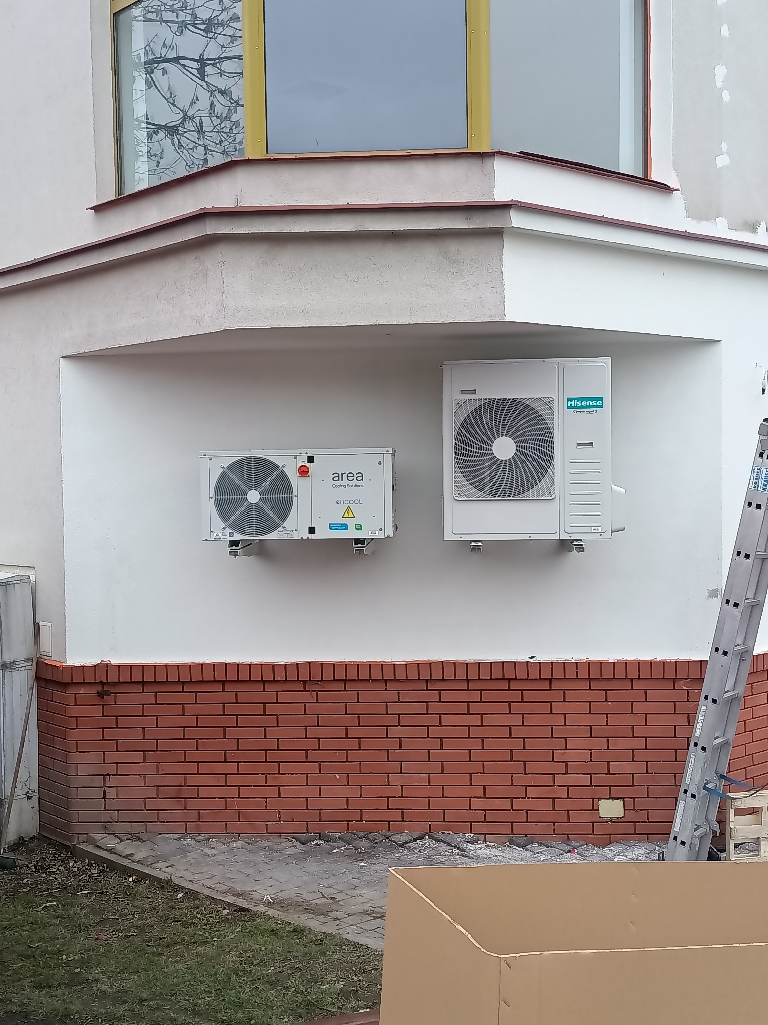 Dwa zamontowane agregaty klimatyzacyjne różnych marek (Area i Hisense) na elewacji budynku z czerwoną cegłą, widoczna drabina oparta o ścianę i kartonowe pudło na pierwszym planie.