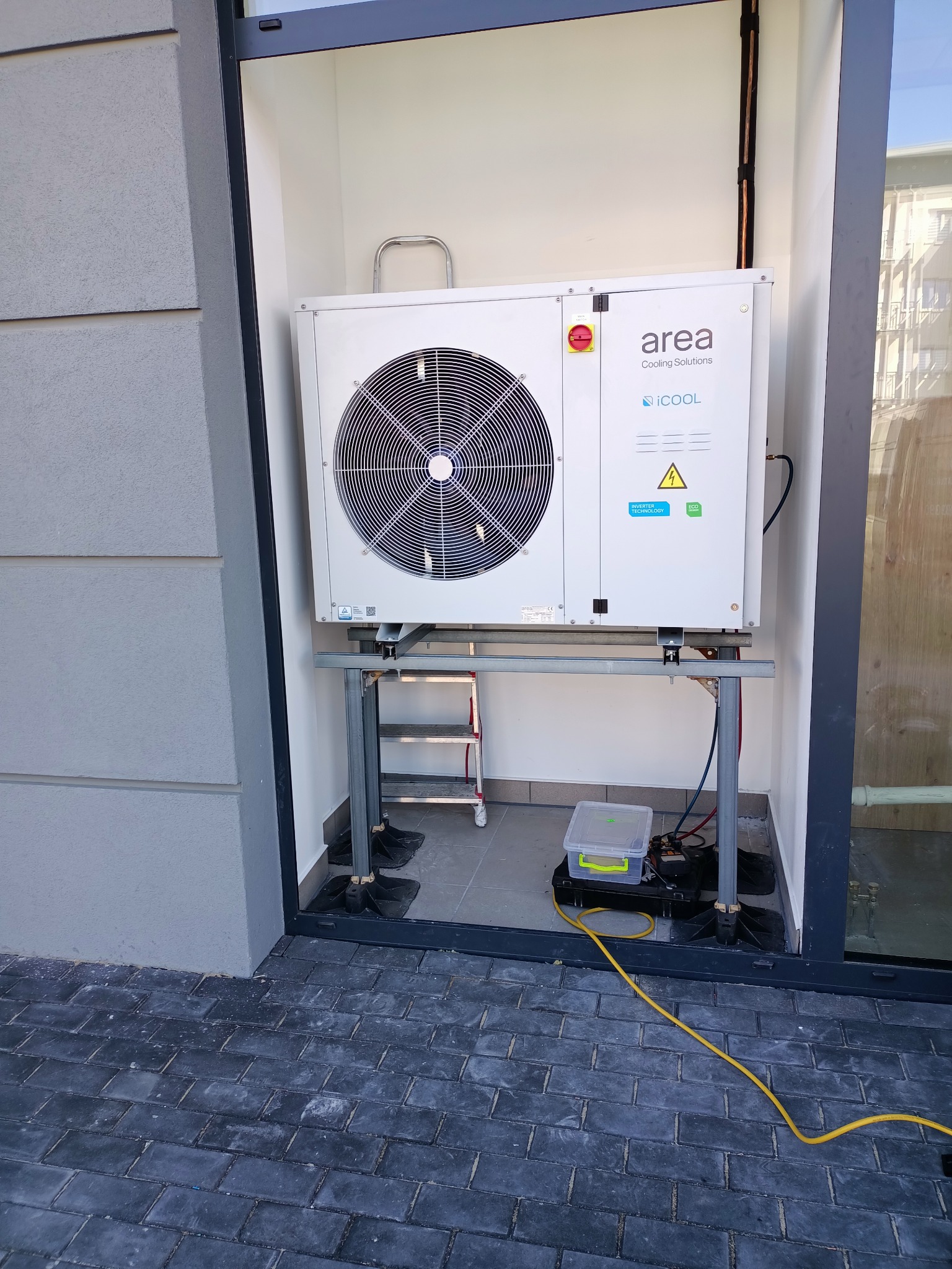 Zewnętrzna jednostka klimatyzacji Area Cooling Solutions iCOOL zamontowana na metalowej konstrukcji, widoczna drabinka i narzędzia serwisowe w pobliżu, instalacja na zewnątrz budynku.