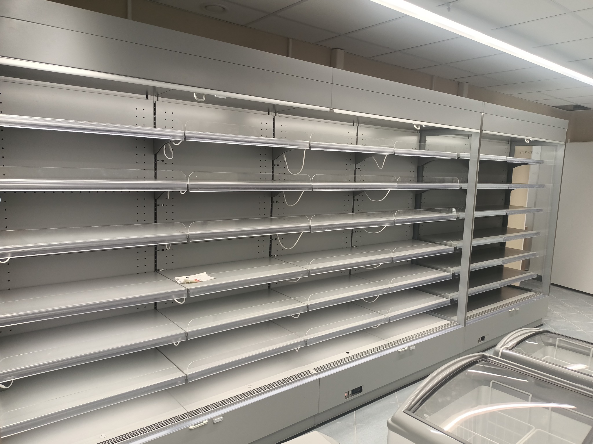 Puste, szare regały chłodnicze w sklepie w Radomiu, widoczne przewody i uchwyty na półkach, z przodu przeszklone lady chłodnicze.