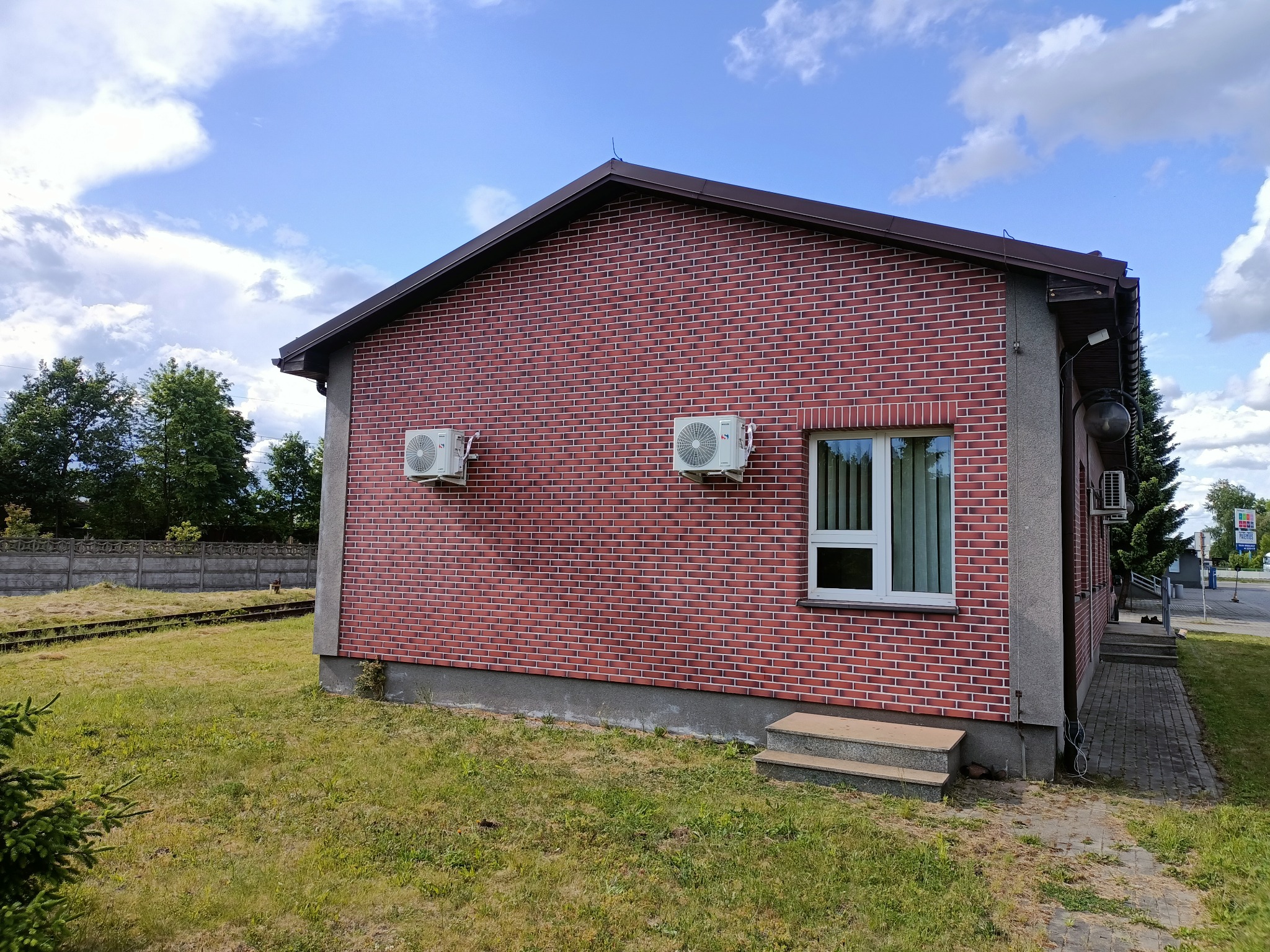 Dwa klimatyzatory zamontowane na ceglanej ścianie budynku z brązowym dachem, obok okna z białymi roletami, widok z zewnątrz na trawnik i fragment torów kolejowych w tle.