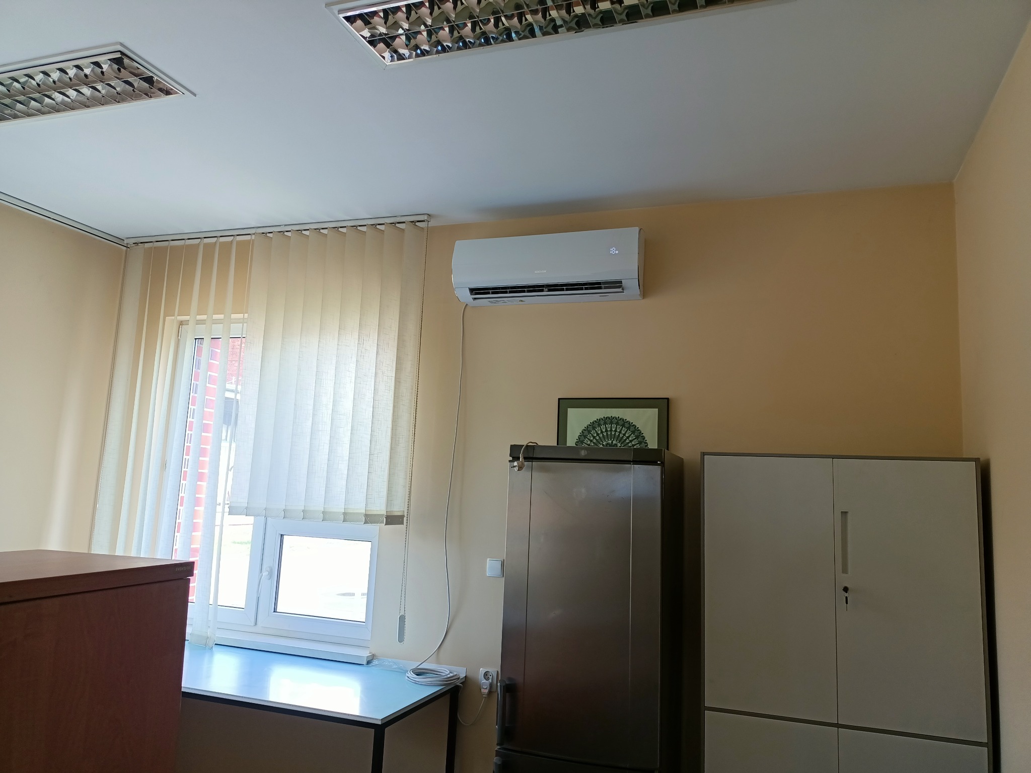 Biały klimatyzator ścienny zamontowany w biurze nad metalową szafką, widoczne okno z żaluzjami pionowymi i biurko.