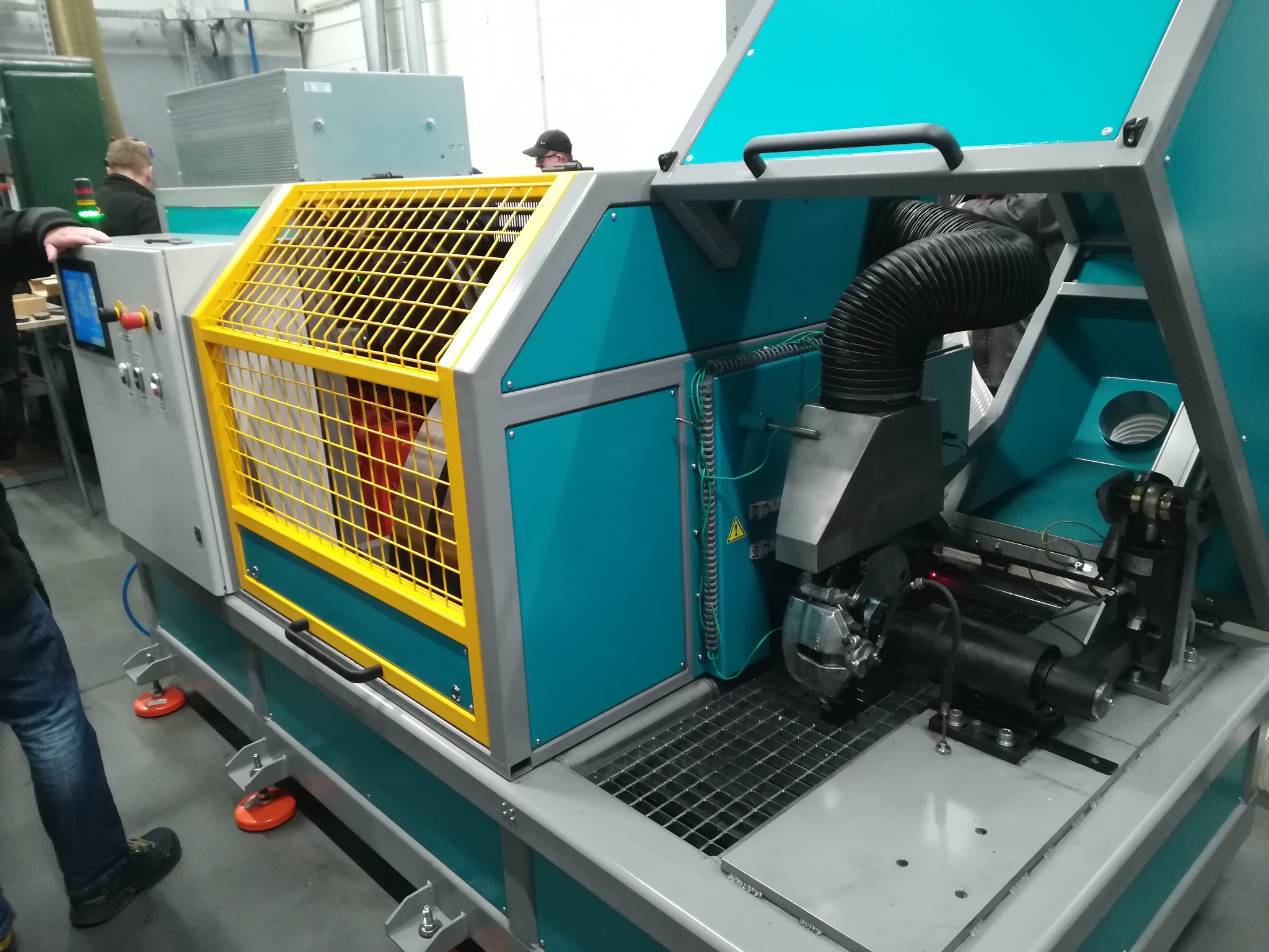 Nowoczesna maszyna przemysłowa z elementami wentylacji, obudowana w turkusowo-szarą konstrukcję z żółtą kratą zabezpieczającą, widoczny fragment osoby obsługującej panel sterowania.
