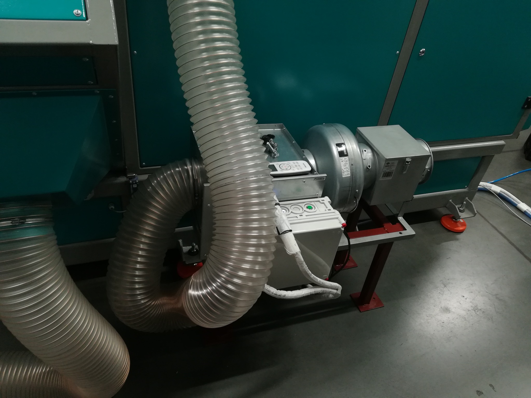 Fragment instalacji wentylacyjnej z widocznymi elastycznymi przewodami, wentylatorem i skrzynką sterowniczą z przyciskami na tle zielonej obudowy urządzenia.