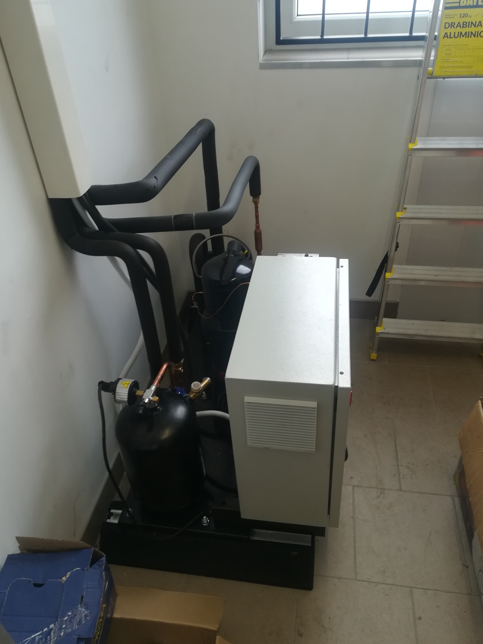 Zainstalowany system klimatyzacji z czarnymi rurami, białym panelem sterowania i aluminiową drabiną w rogu pomieszczenia.