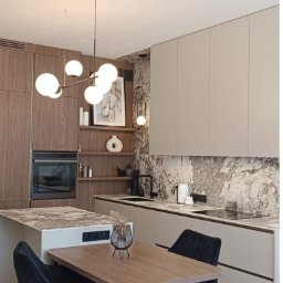 s'Kubana Anna Skrobisz-Bujanowska - Nowoczesna kuchnia z drewnianymi i beżowymi szafkami, marmurowym backsplashiem, wyspą z blatem imitującym kamień, okrągłym żyrandolem i stołem z czarnymi krzesłami obrotowymi.