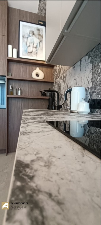 Nowoczesna kuchnia z blatem imitującym marmur, czarną armaturą i białym czajnikiem elektrycznym, uzupełniona o drewniane półki z dekoracjami i grafiką.