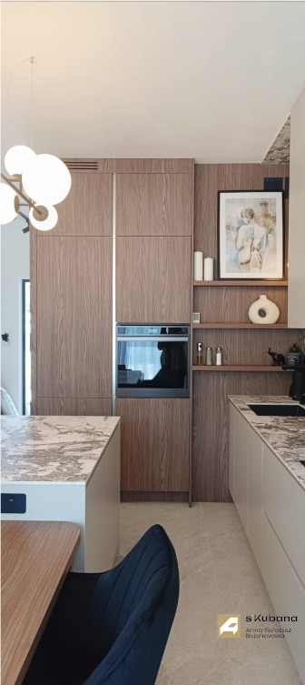 Nowoczesna kuchnia z drewnianymi frontami, marmurowym blatem i wbudowanym piekarnikiem, uzupełniona dekoracjami na półkach i designerską lampą.