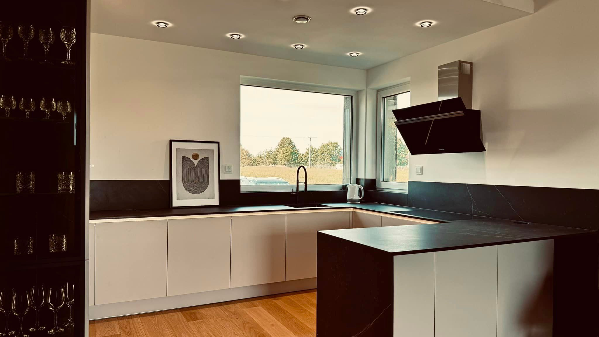 Nowoczesna kuchnia z jasnymi szafkami dolnymi, blatem z ciemnego kamienia, czarną armaturą i oknem z widokiem na zieleń. Na blacie czajnik elektryczny, nad zlewem minimalistyczna grafika w ramie,...