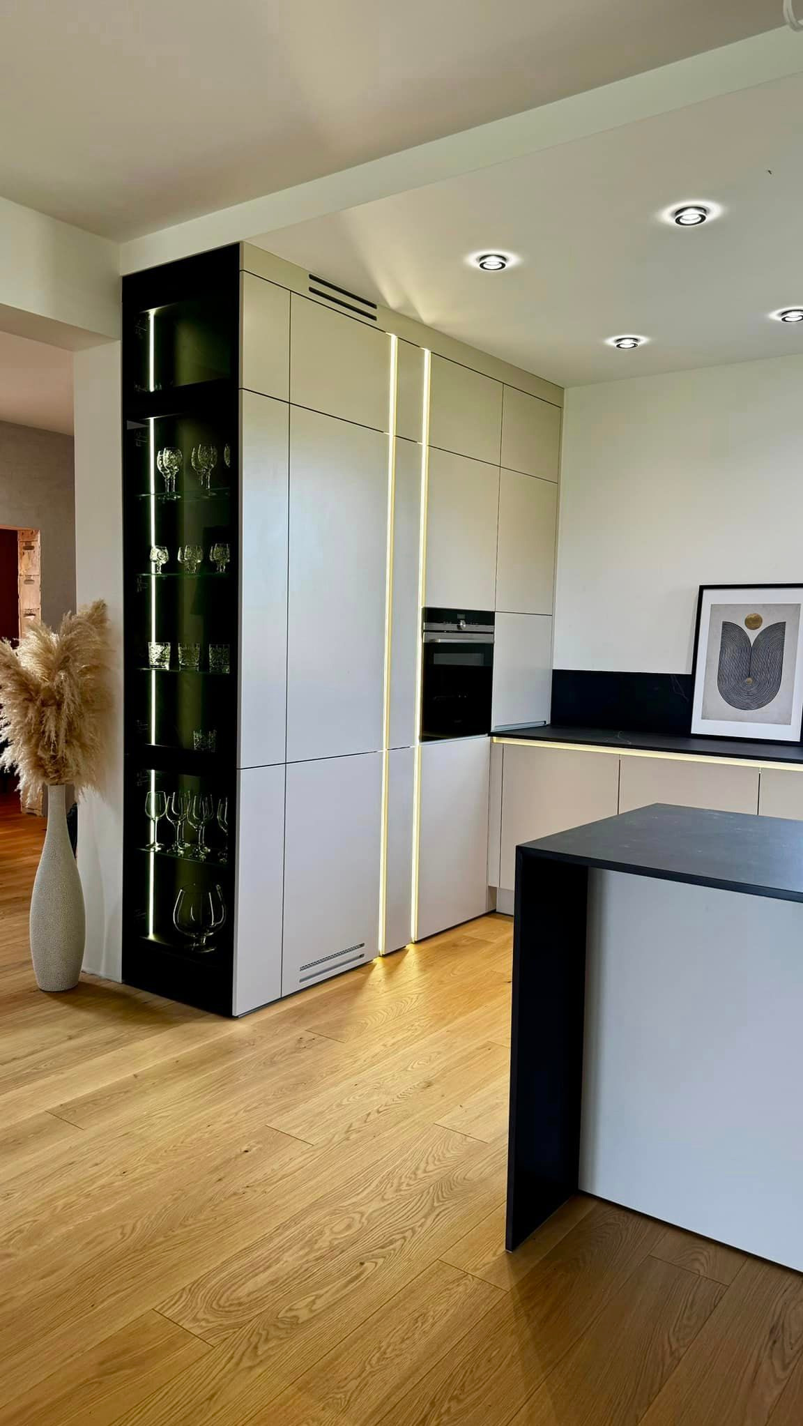 Nowoczesna kuchnia z zabudową meblową w odcieniach bieli i beżu, z oświetleniem LED w pionowych szczelinach między szafkami i przeszkloną witryną z kieliszkami, na drewnianej podłodze.