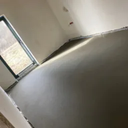 Wylany, świeży jastrych w pomieszczeniu z widocznymi przewodami elektrycznymi na ścianie i listwami dylatacyjnymi przy ścianach oraz oknem z widokiem na zewnątrz.