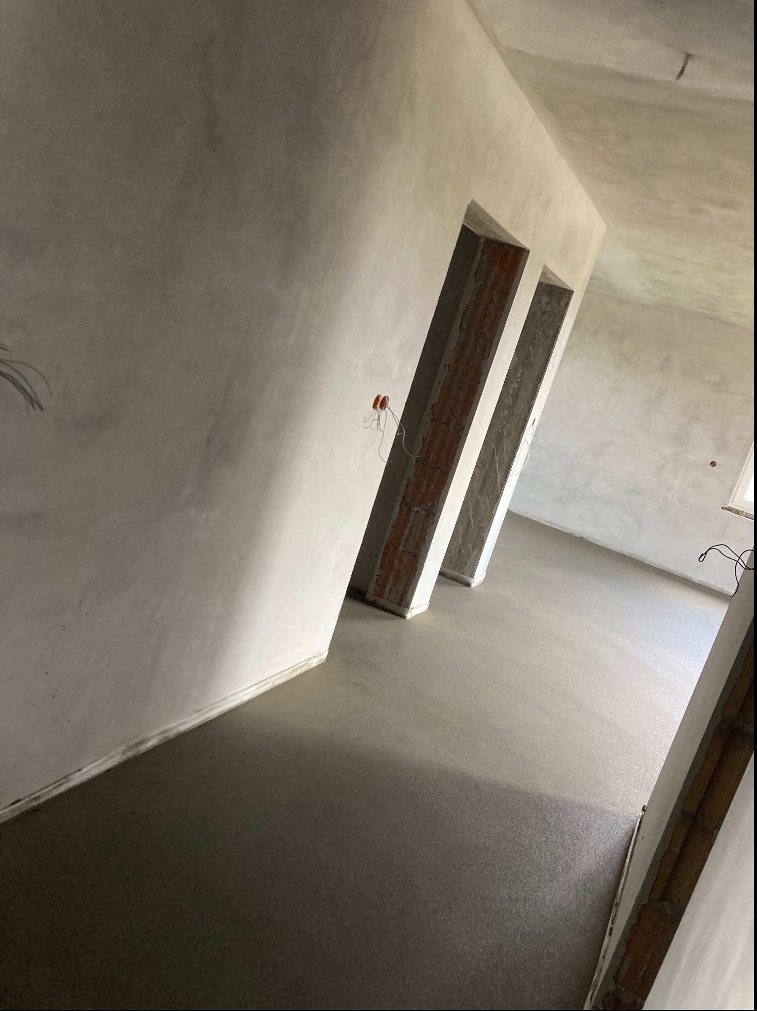 Świeżo wylana, gładka posadzka w jasnym pomieszczeniu z widocznymi, surowymi framugami drzwi i przewodami elektrycznymi na ścianie.