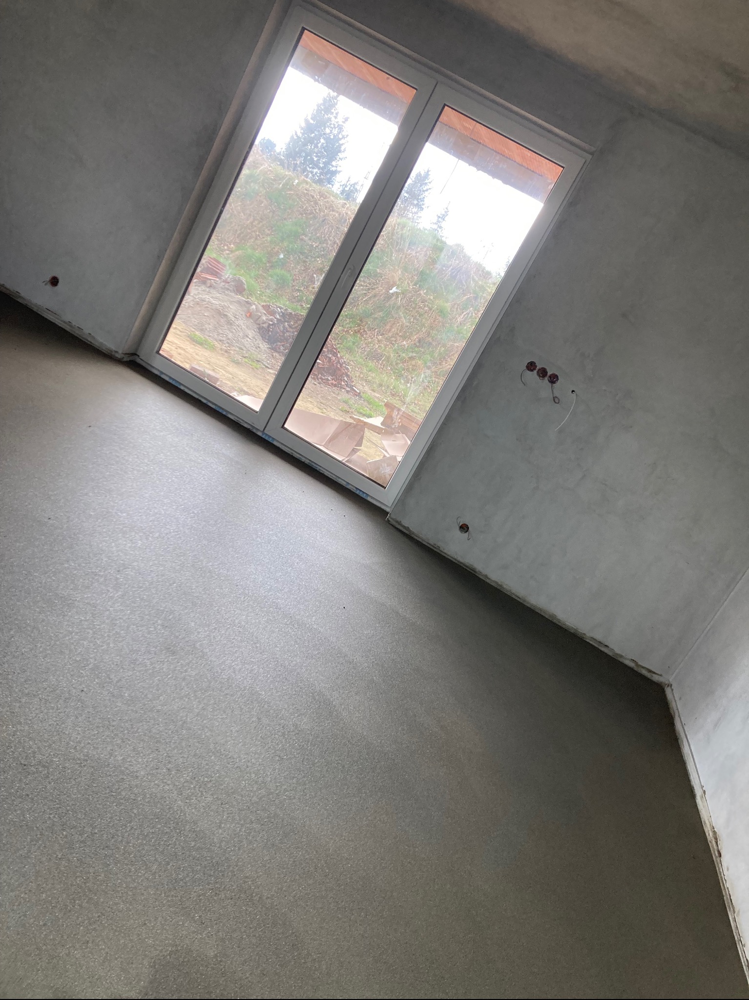 Świeżo wylana, gładka posadzka w surowym wnętrzu z dużym oknem balkonowym, przez które widać zewnętrzny krajobraz z ziemią i roślinnością.