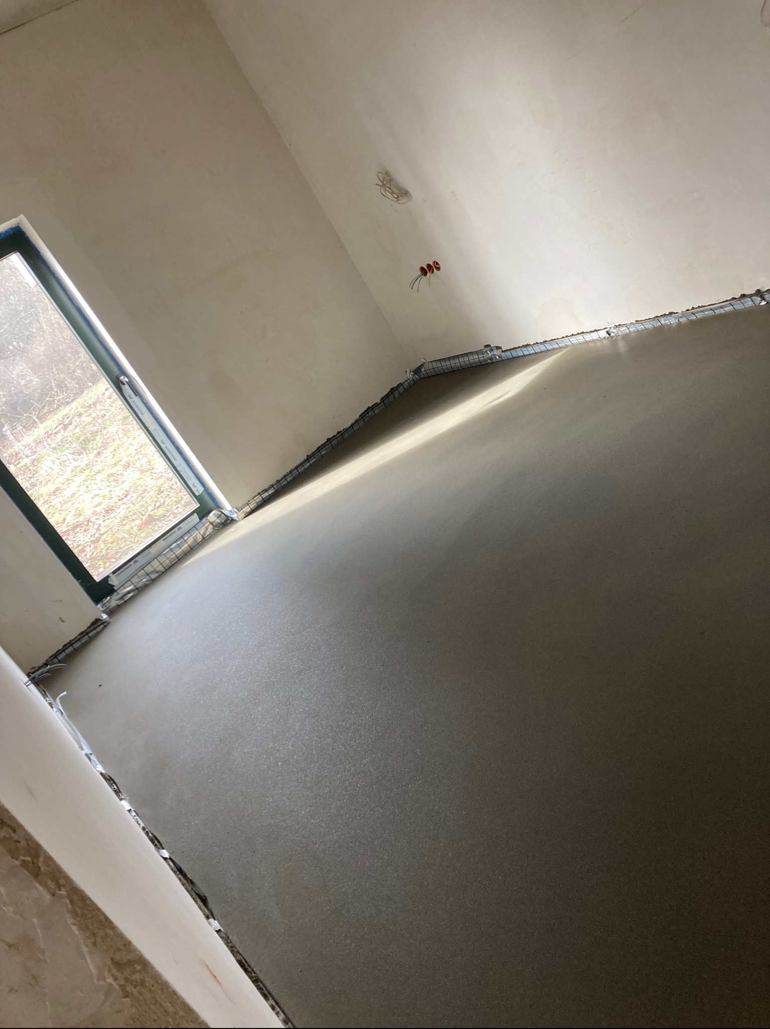 Wylany, świeży jastrych w pomieszczeniu z widocznymi przewodami elektrycznymi na ścianie i listwami dylatacyjnymi przy ścianach oraz oknem z widokiem na zewnątrz.