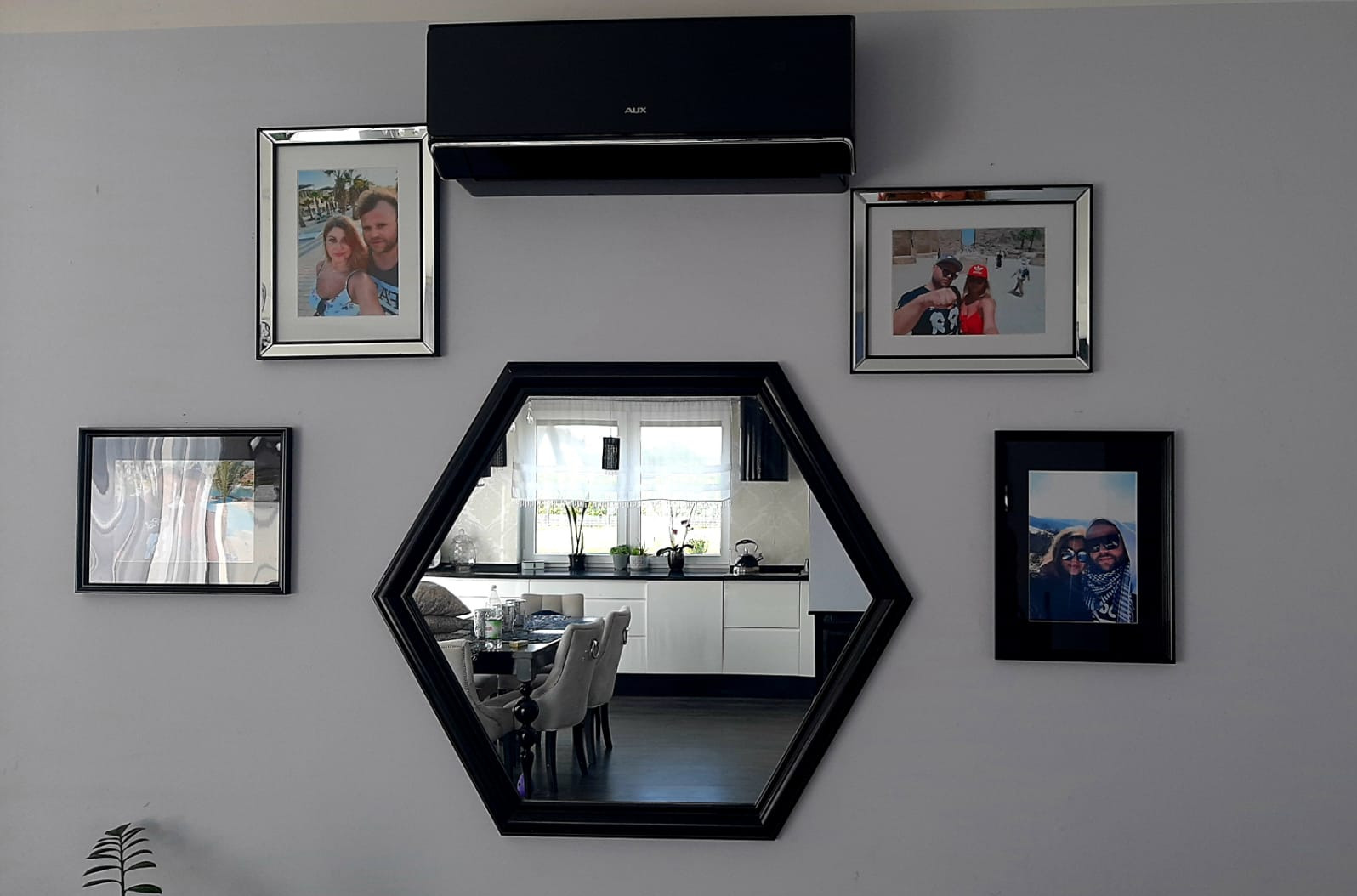 Czarna jednostka klimatyzacji AUX zamontowana na szarej ścianie nad heksagonalnym lustrem i ramkami ze zdjęciami rodzinnymi.