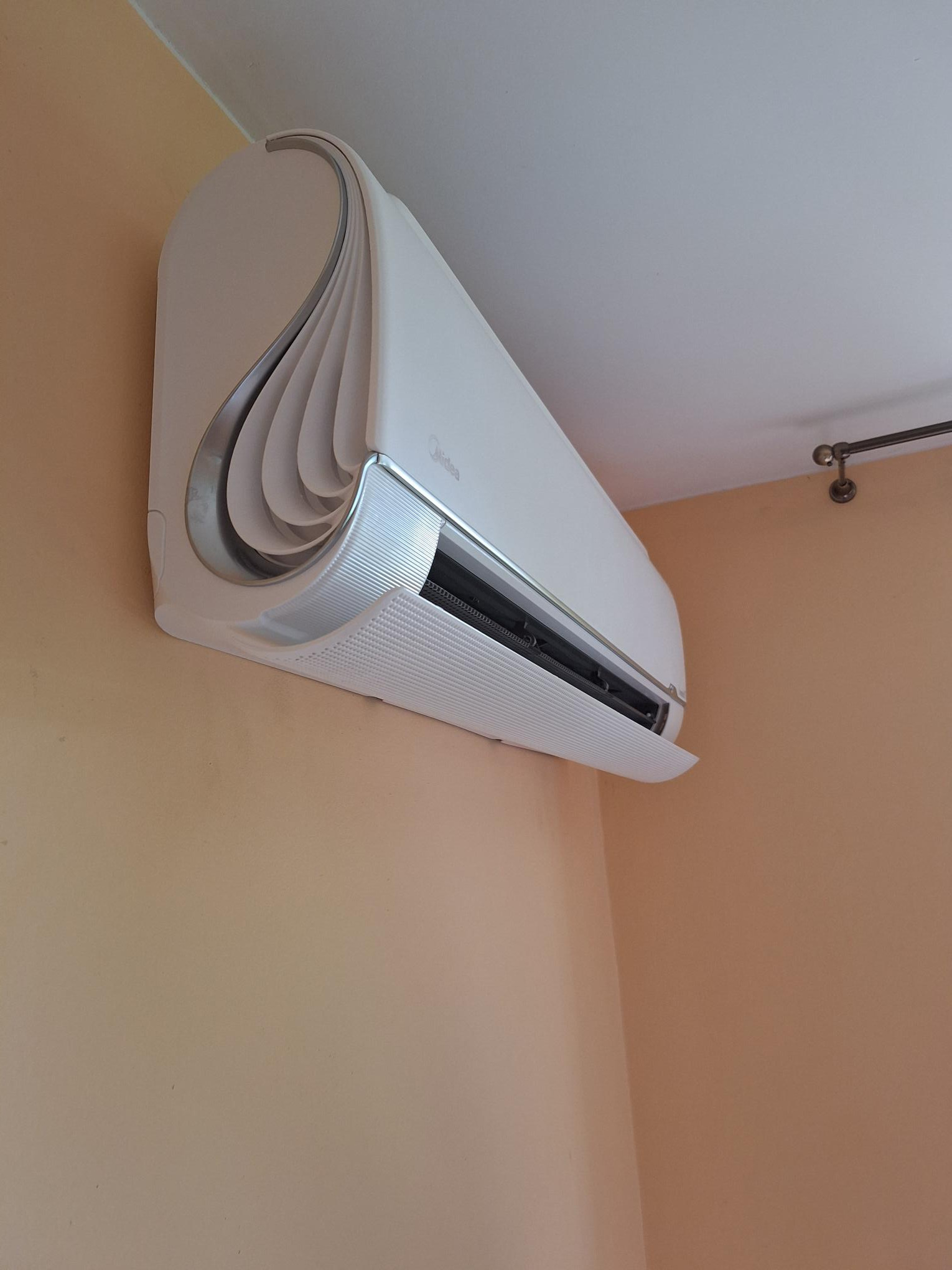 Nowoczesny, biały klimatyzator Midea zamontowany na ścianie o brzoskwiniowym odcieniu, widoczny fragment sufitu i karnisz.