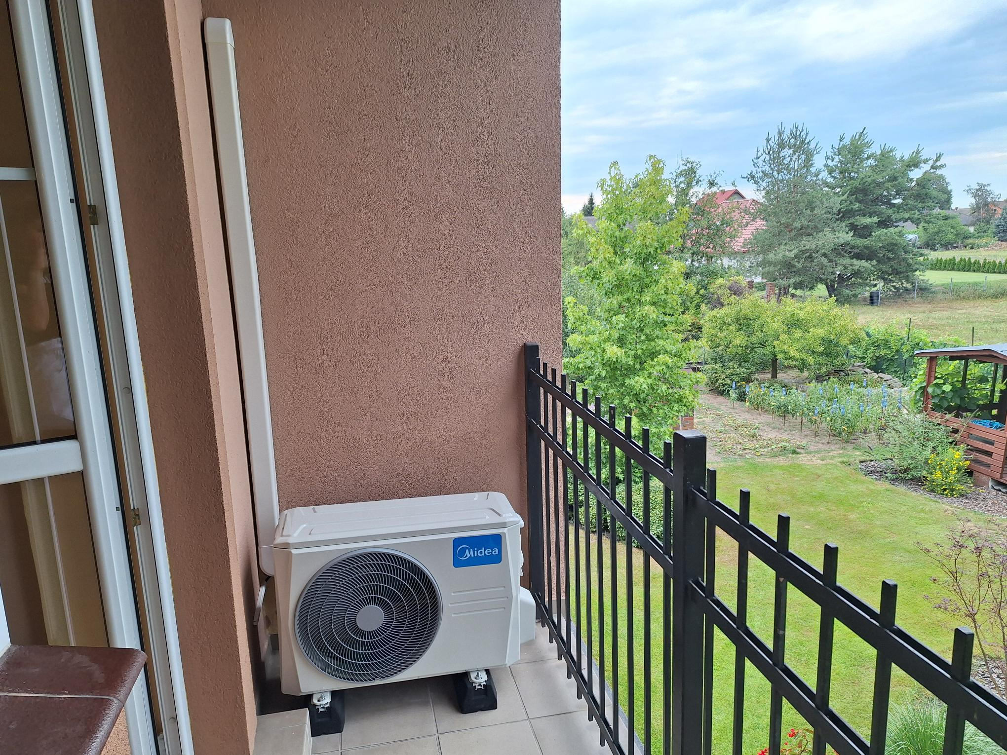 Zewnętrzna jednostka klimatyzacji marki Midea zamontowana na balkonie z widokiem na zielony ogród.