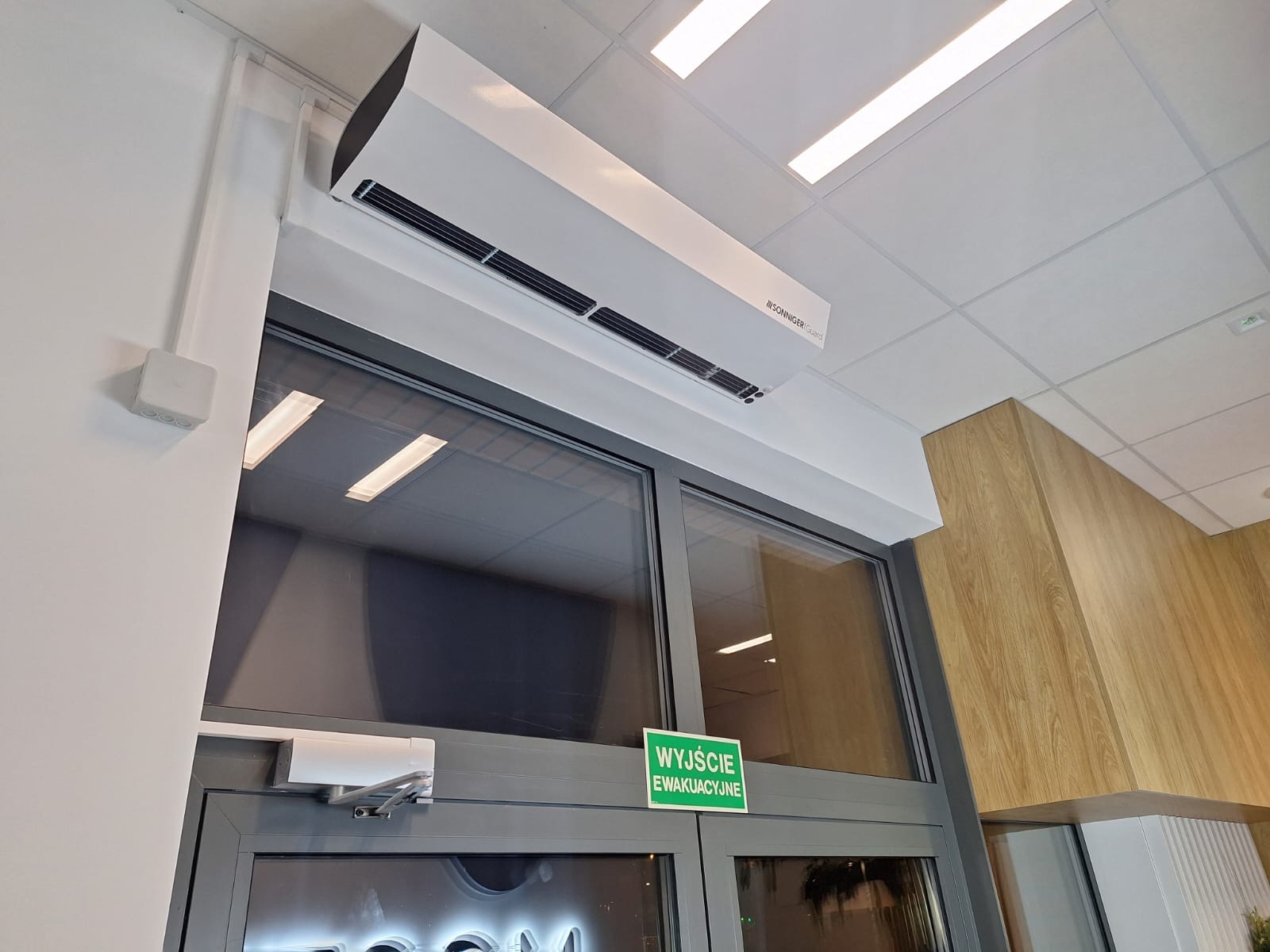 Biała kurtyna powietrzna marki Sonniger Guard zamontowana nad szarymi drzwiami ewakuacyjnymi z zielonym znakiem, widok z dołu na sufit kasetonowy i drewnianą okładzinę ściany.
