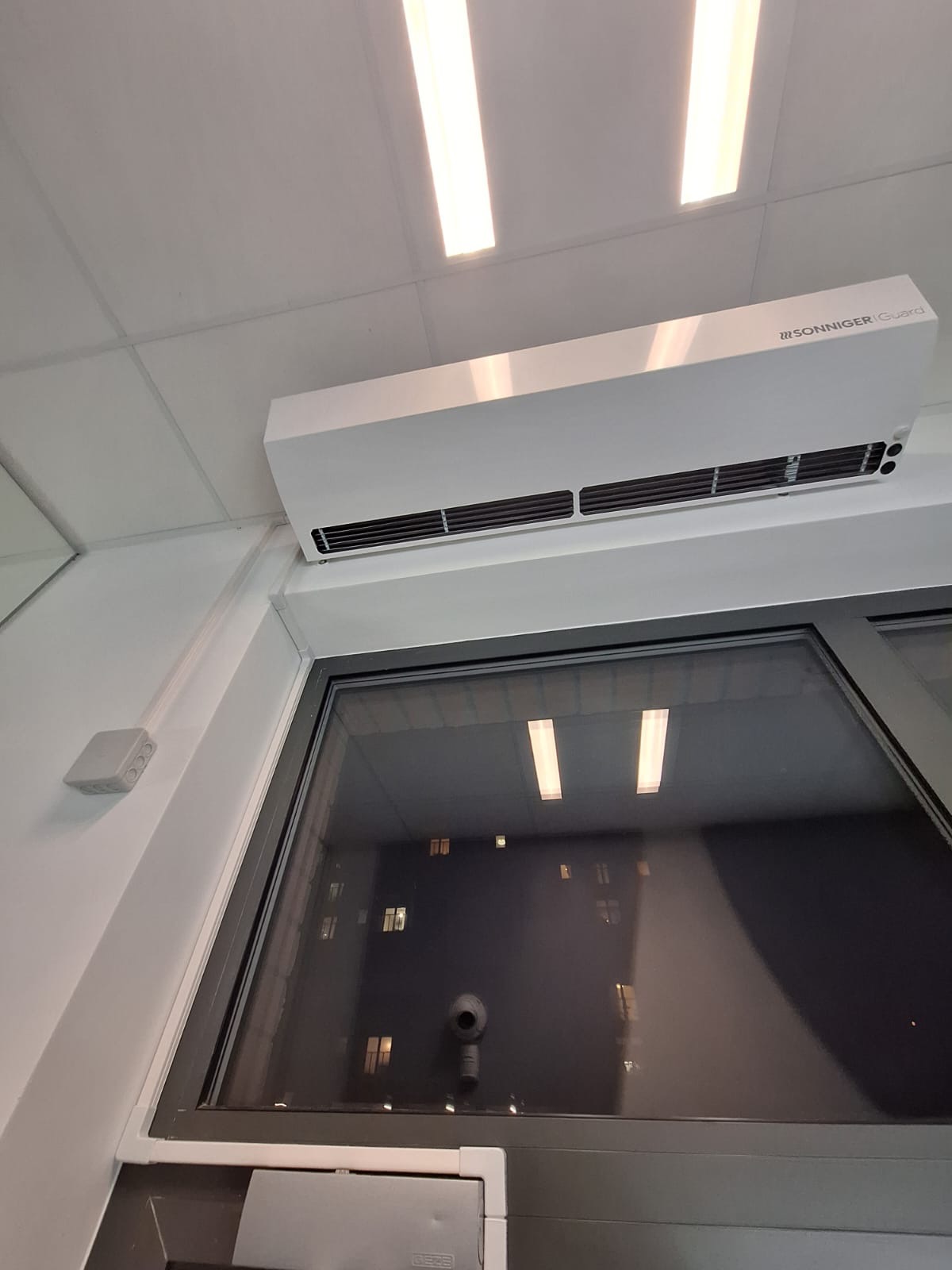 Biała jednostka klimatyzacyjna Sonniger Guard zamontowana pod sufitem w biurze, widoczne okno z odbiciem świateł budynku.