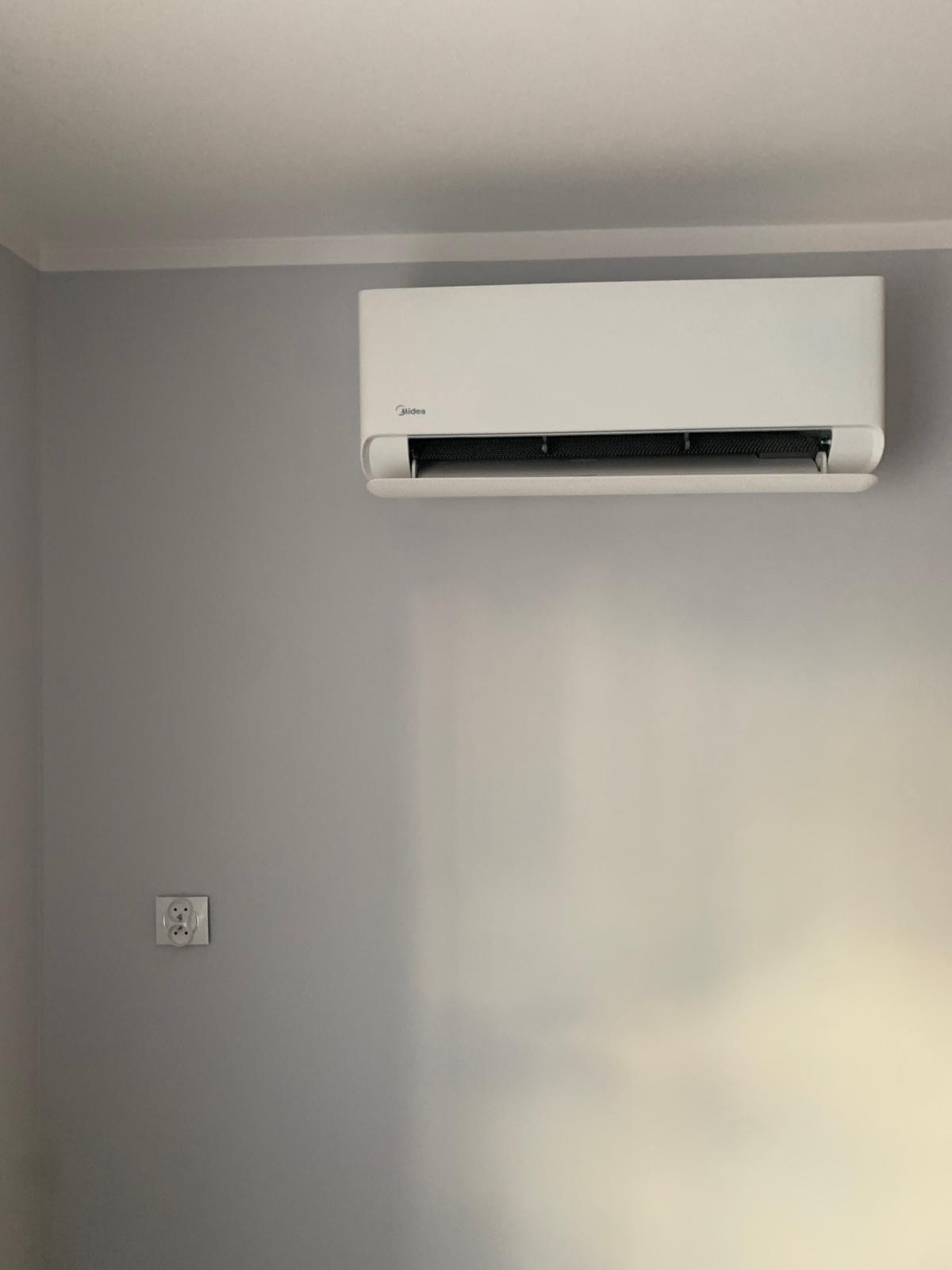 Biały klimatyzator Midea zamontowany na szarej ścianie wewnątrz pomieszczenia, widoczne gniazdko elektryczne po lewej stronie.
