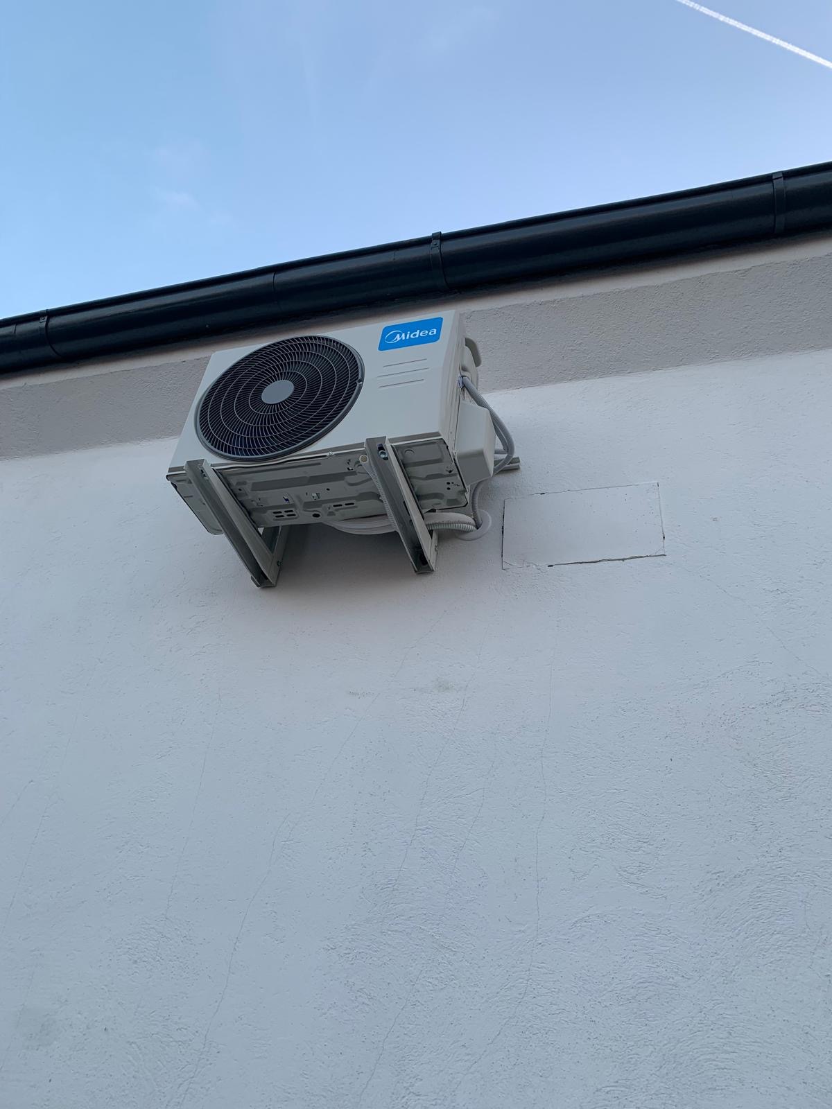 Zewnętrzna jednostka klimatyzacji Midea zamontowana na białej ścianie budynku, widoczne metalowe wsporniki i przewody, zarys nieba w tle.