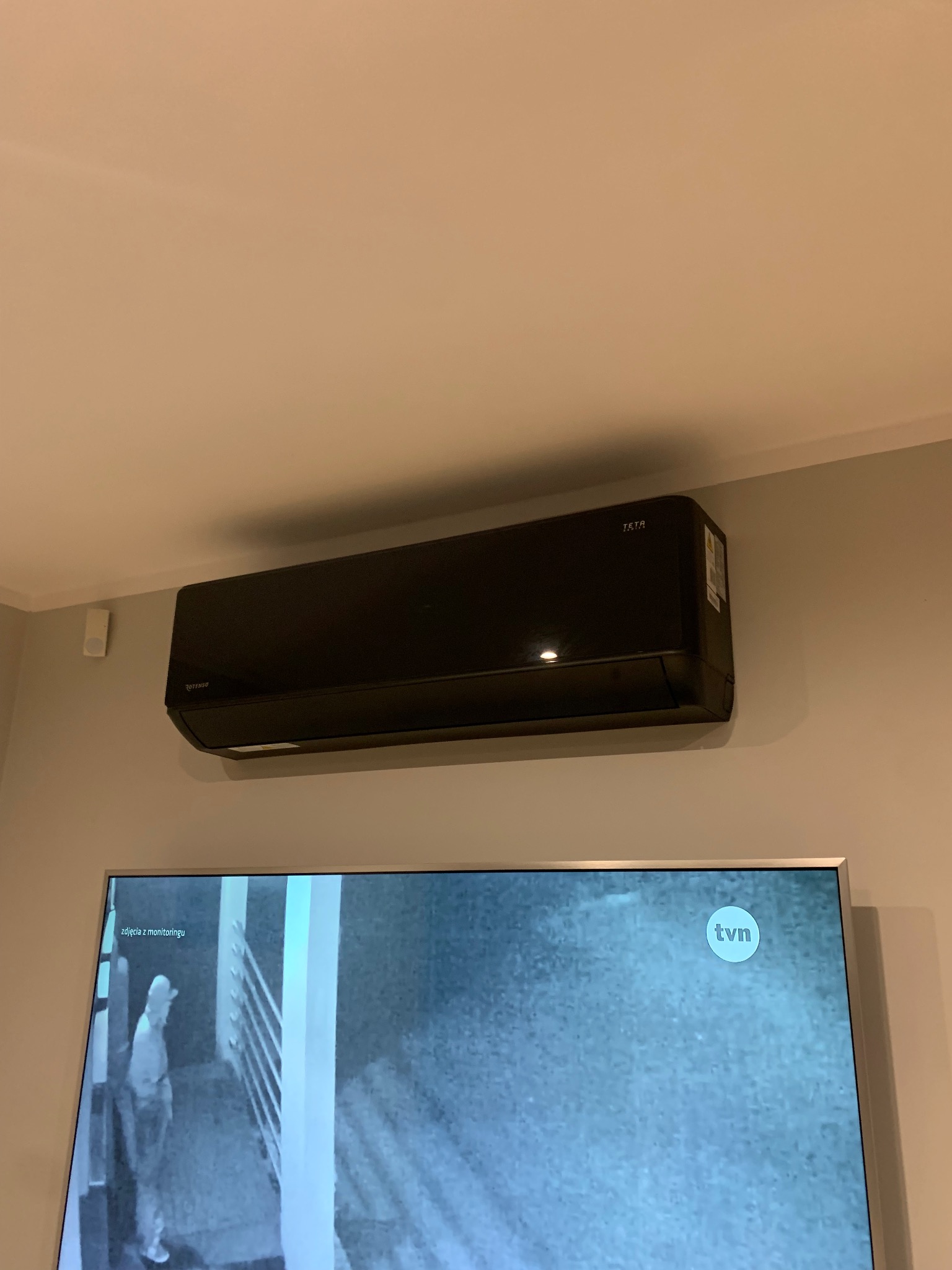 Czarny klimatyzator marki Rotenso zamontowany na ścianie nad telewizorem z widocznym fragmentem obrazu z monitoringu.