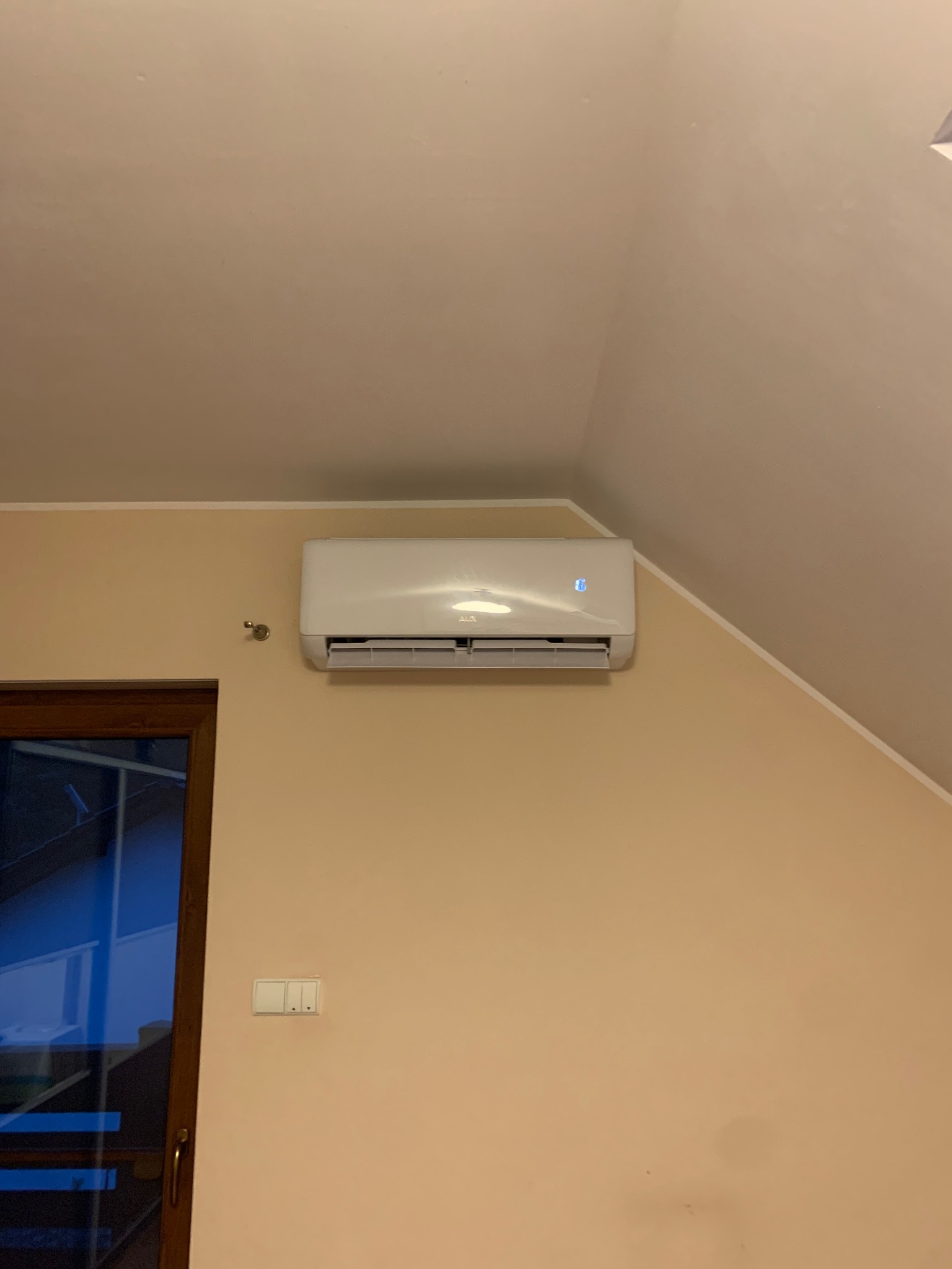 Biała jednostka klimatyzacyjna zamontowana na beżowej ścianie w pokoju na poddaszu, widoczny fragment okna i włączniki światła.