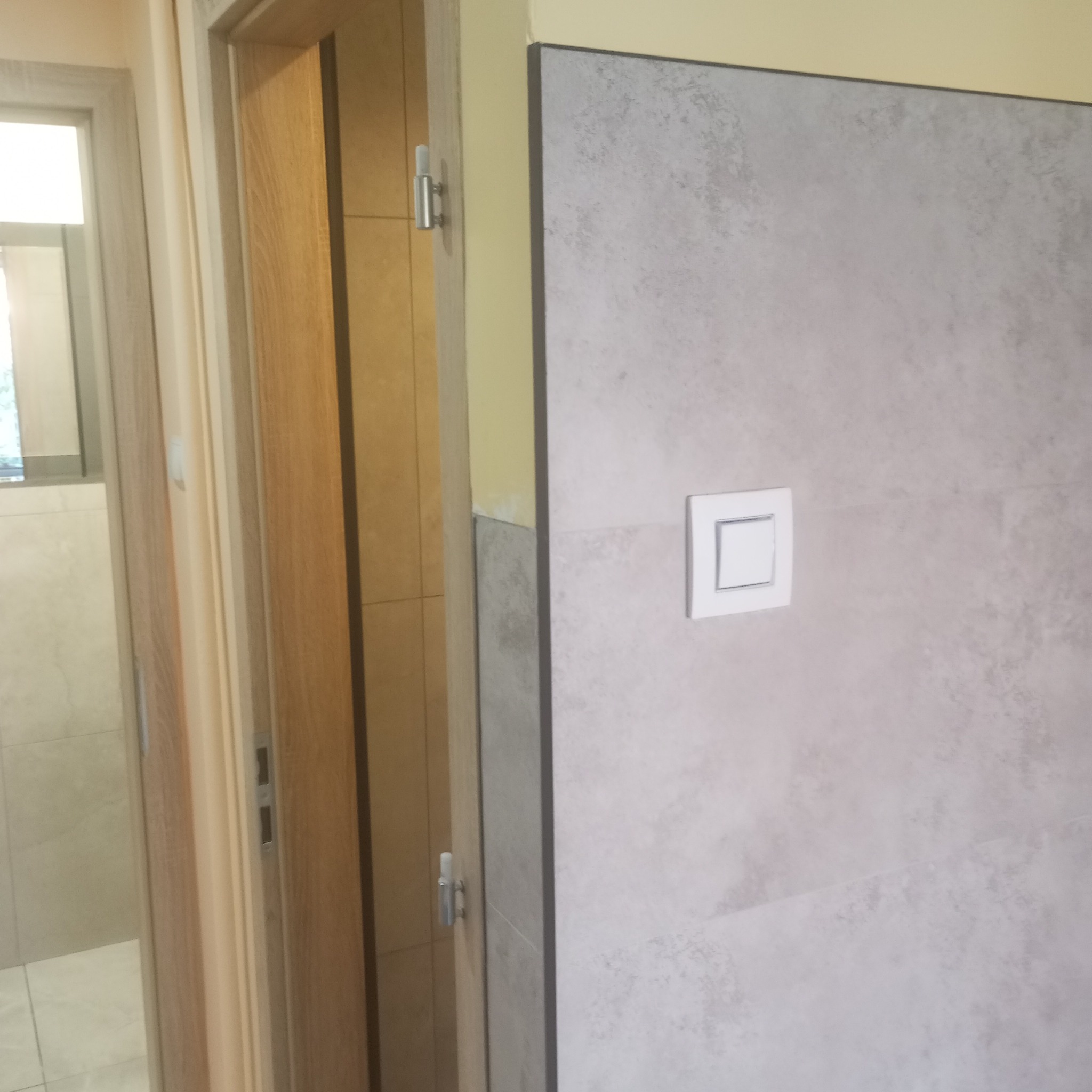 Wykończona ściana w łazience z szarymi płytkami imitującymi kamień i białym włącznikiem światła. Widoczne fragmenty dwóch ościeżnic drzwiowych.