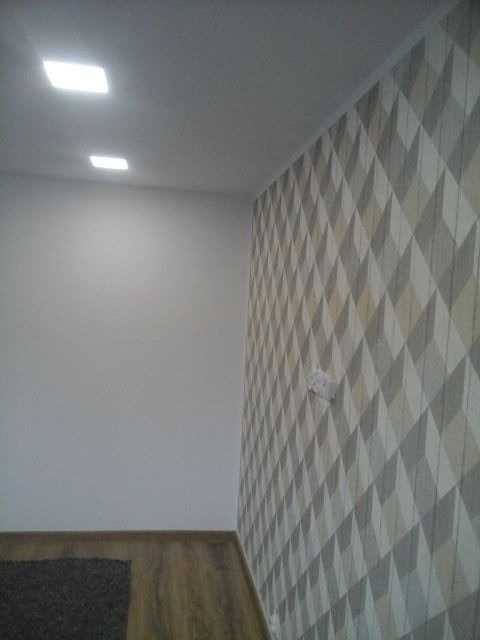 Wykończone wnętrze: ściana z geometryczną tapetą w odcieniach szarości i beżu, kontrastująca z gładką, białą ścianą. Widoczne panele podłogowe i minimalistyczne oświetlenie sufitowe.