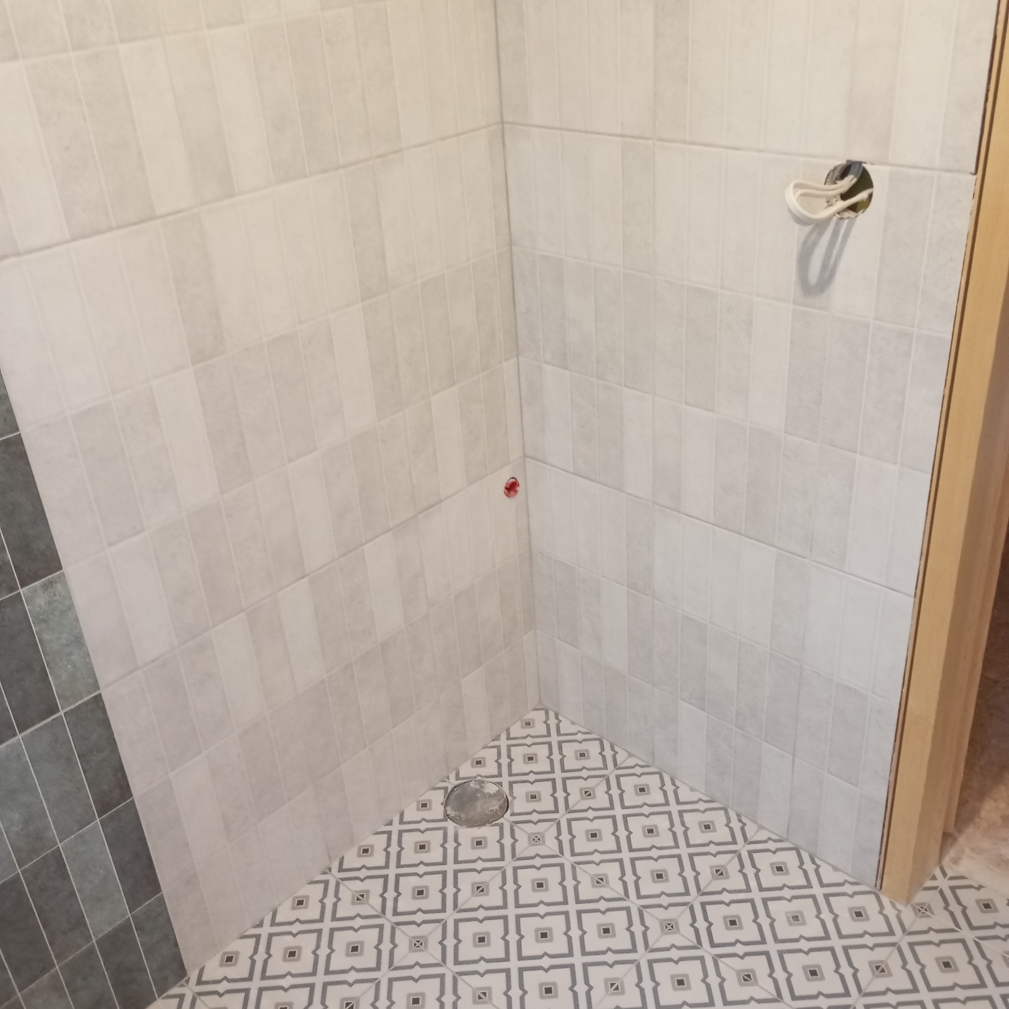 Wykończona łazienka z jasnymi, pionowo ułożonymi płytkami na ścianach i wzorzystą podłogą. Widoczne gniazdko elektryczne i fragment drzwi.