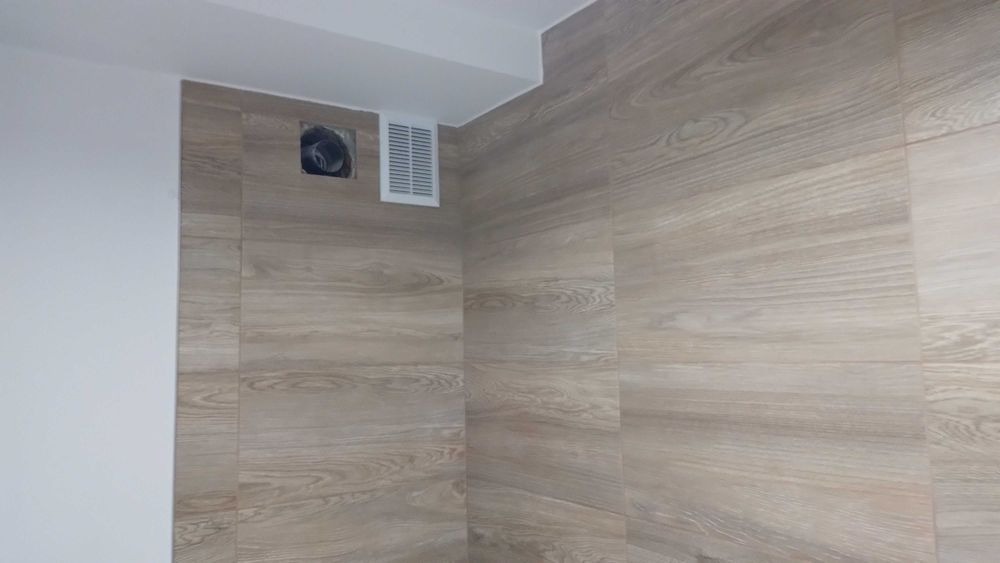 Ściana wykończona płytkami imitującymi drewno, z widoczną kratką wentylacyjną i otworem na wentylator. Minimalistyczny design, jasne kolory, nowoczesne wykończenie.