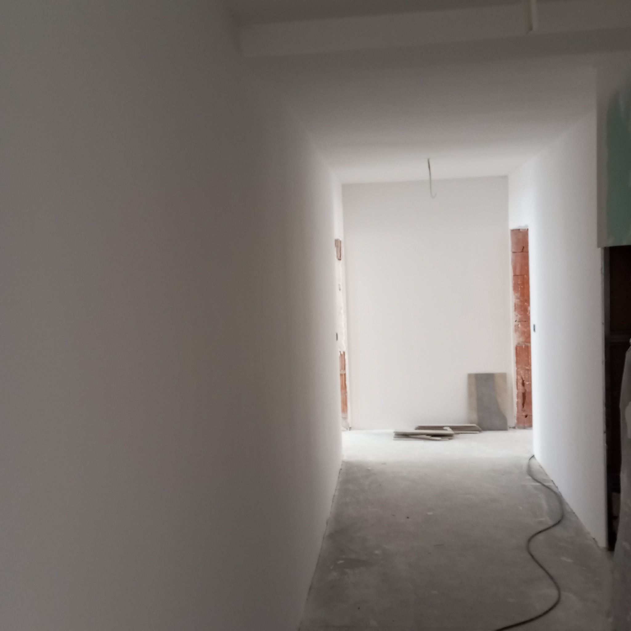 Białe, świeżo pomalowane ściany korytarza w trakcie remontu. Widoczne fragmenty cegieł w otworach drzwiowych i szarej podłogi. Kabel elektryczny leży na podłodze.