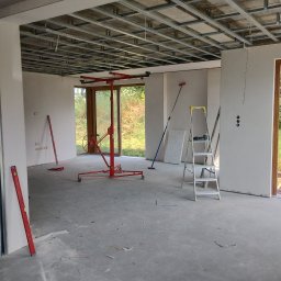 Artur Szulc - Wnętrze w trakcie montażu metalowego rusztu sufitowego podwieszanego, widoczny podnośnik do płyt, poziomice i drabina, surowy betonowy podkład.