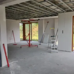 Wnętrze w trakcie montażu metalowego rusztu sufitowego podwieszanego, widoczny podnośnik do płyt, poziomice i drabina, surowy betonowy podkład.